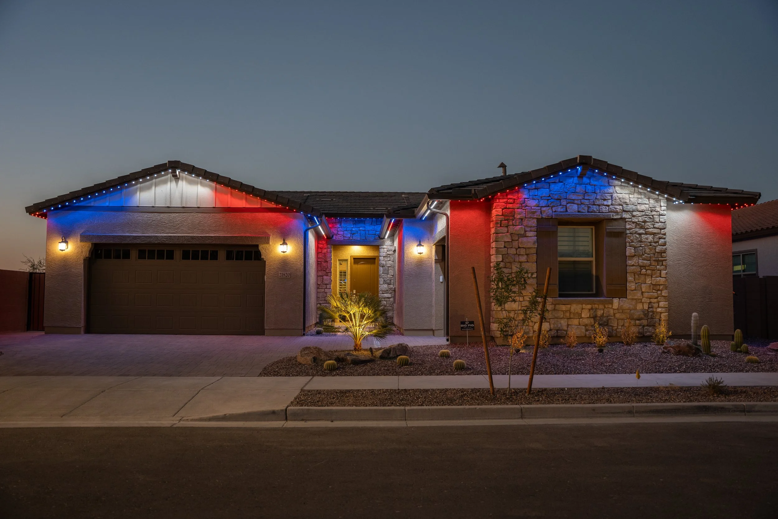 A house decorated with red, white, and blue string lights on the roofline at night. The house has a two-car garage, a front door with a small porch, and desert landscaping with cacti and small bushes.