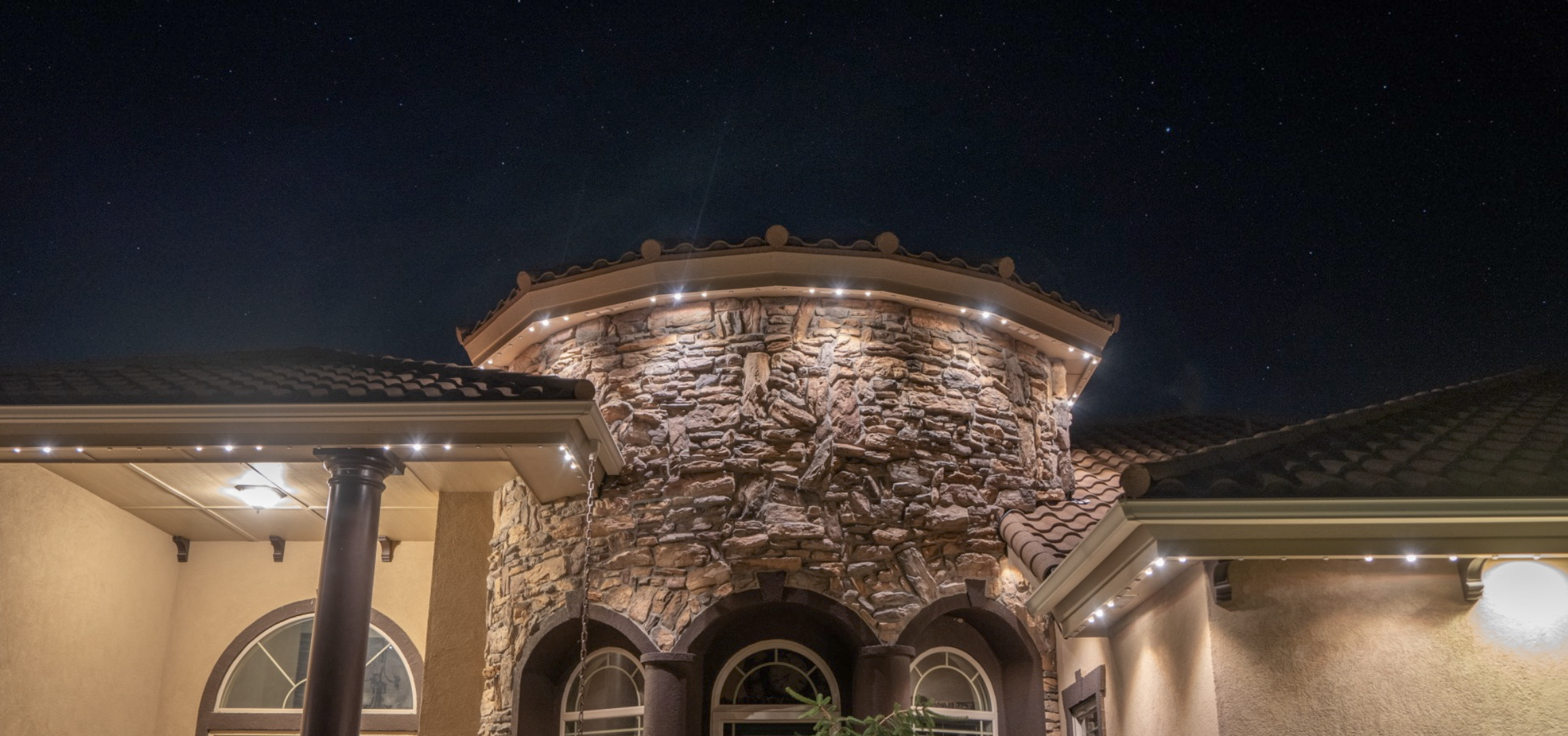 Night view of a house with a stone tower, tile roof, and exterior lights