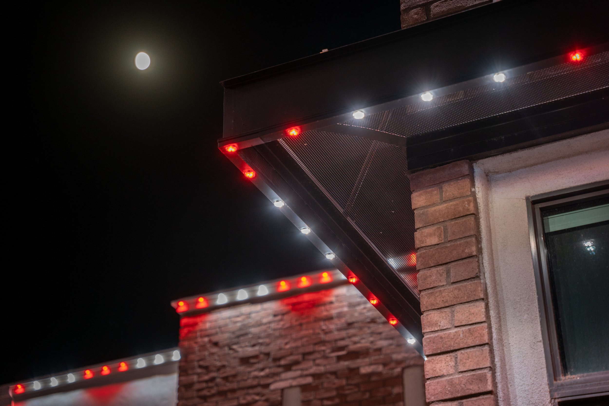 Nighttime view of the exterior corner of a building with brick walls, decorated with white and red outdoor string lights and trim, and a visible window. The sky shows a bright, full moon.