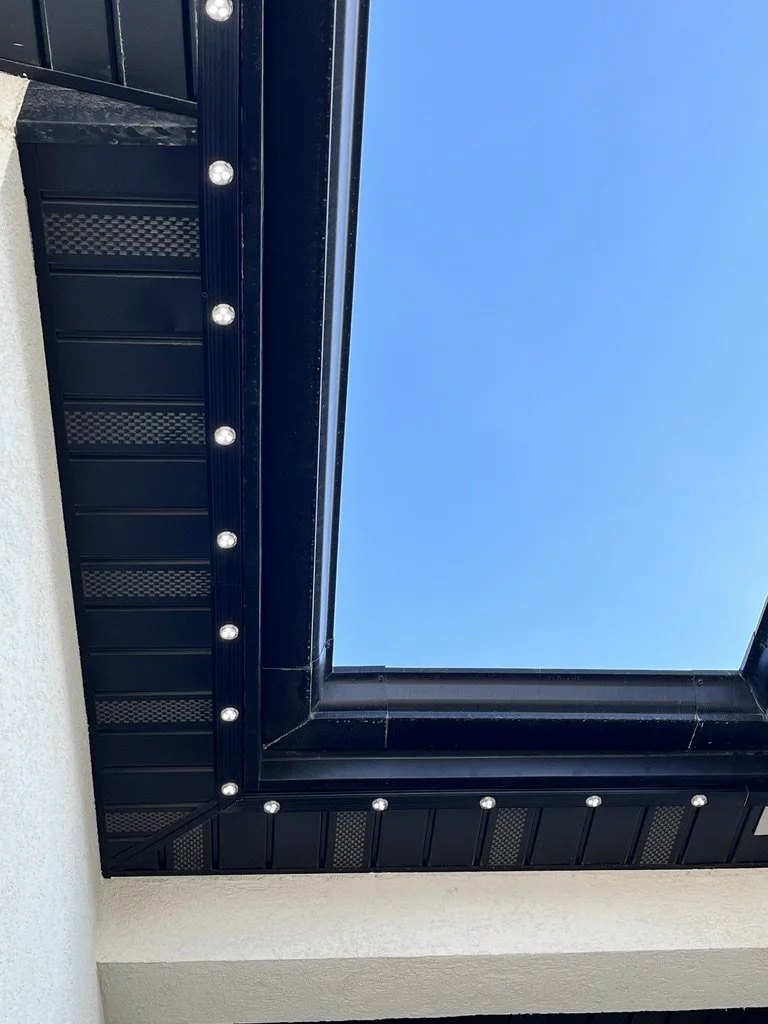 View of a square skylight with a black metal frame, surrounded by a white ceiling and wall, with a clear blue sky visible through the skylight.