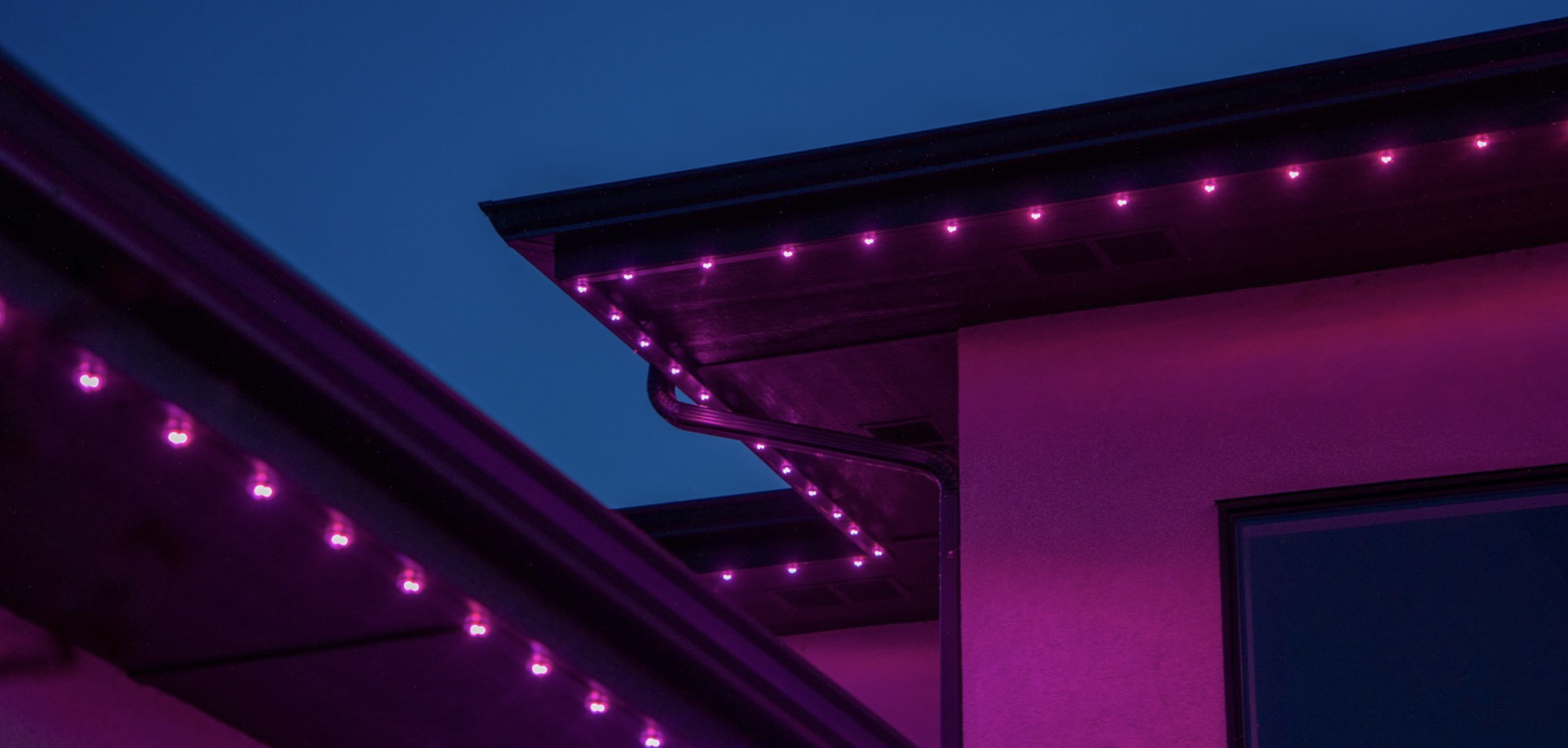 Close-up of a building's roof and wall decorated with purple LED string lights at dusk.