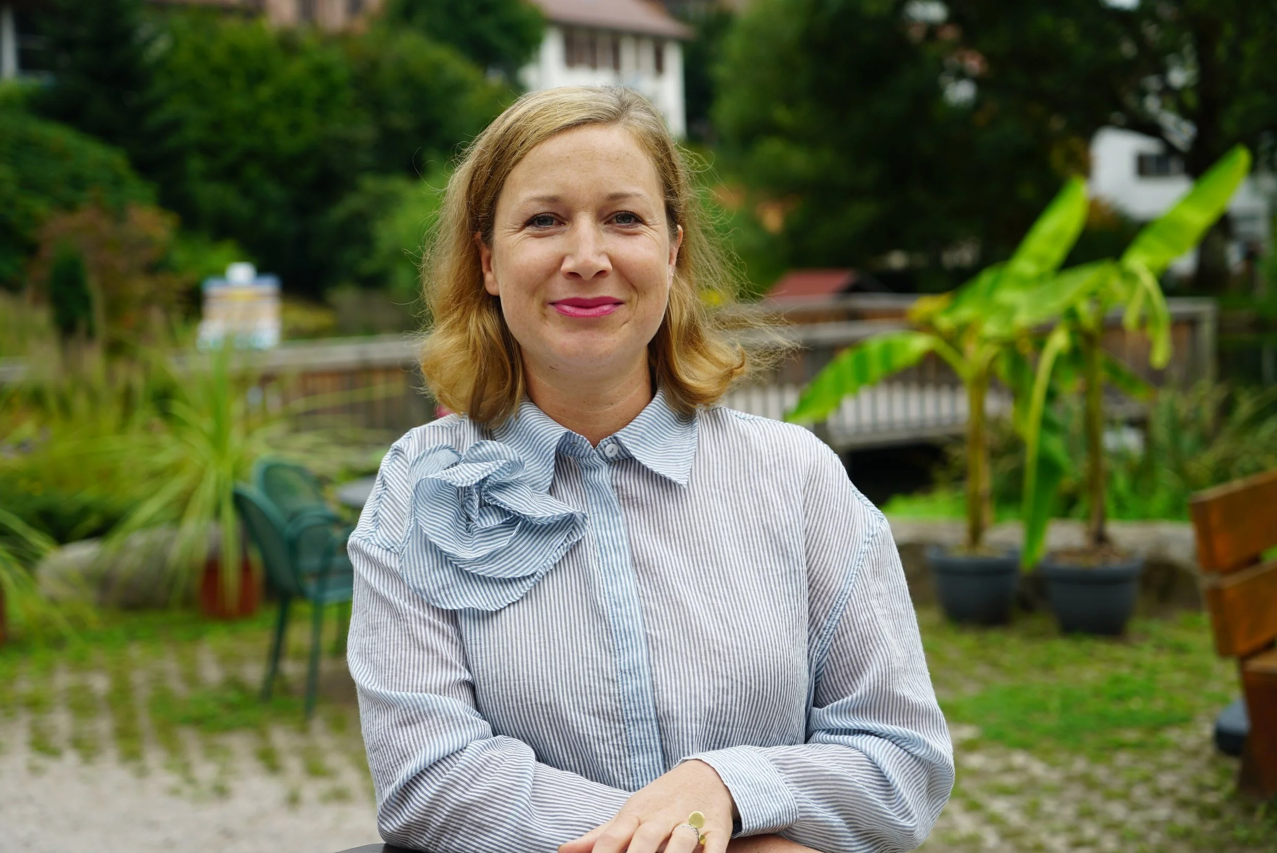 A woman with shoulder-length blonde hair smiling outdoors, wearing a striped blouse with a ruffled collar, in a garden with green plants and a pond in the background.