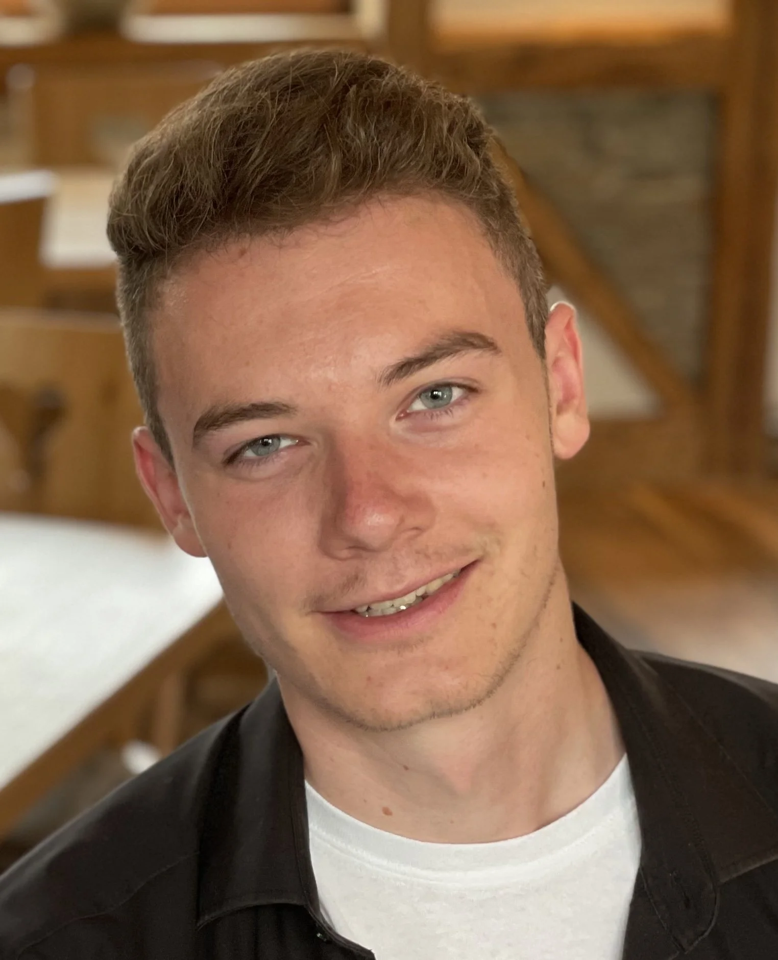 Close-up of a young man with light brown hair, blue eyes, and a fair complexion, smiling at the camera, in an indoor setting with wooden furniture in the background.