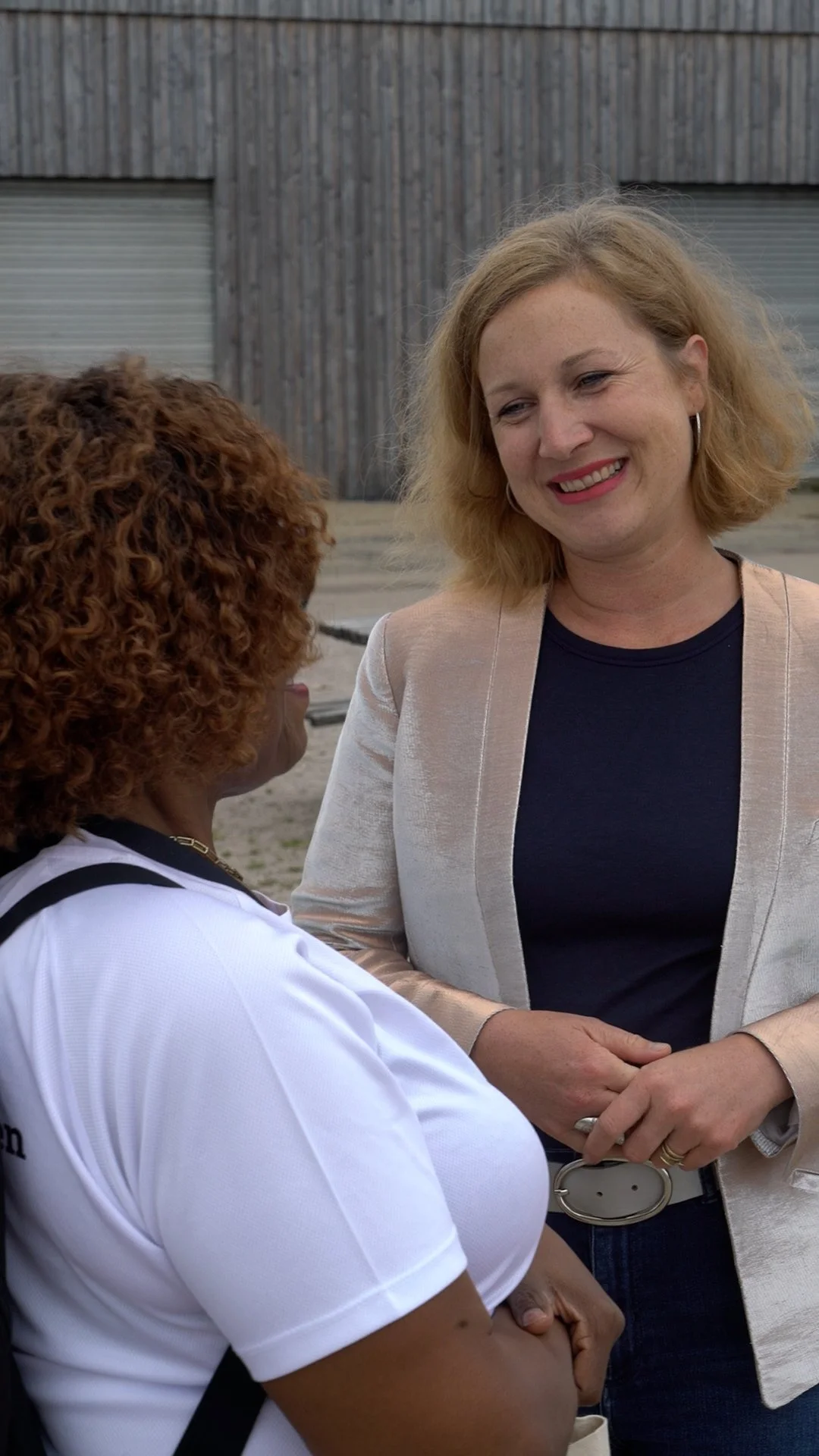 Two women talking outdoors, one with curly red hair wearing a white shirt, the other with blonde hair wearing a beige blazer over a black shirt.