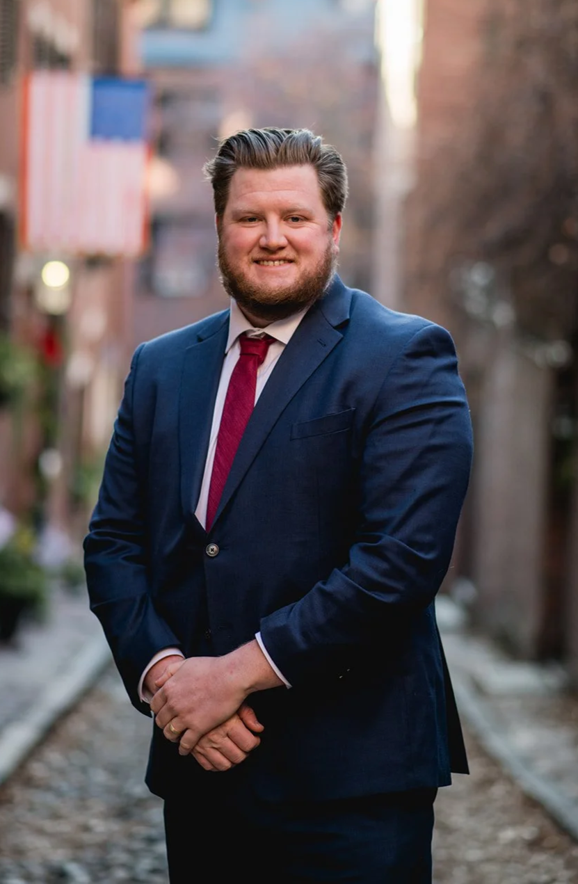 A man in a dark blue suit, white shirt, and red tie standing outdoors on a city street, smiling at the camera.