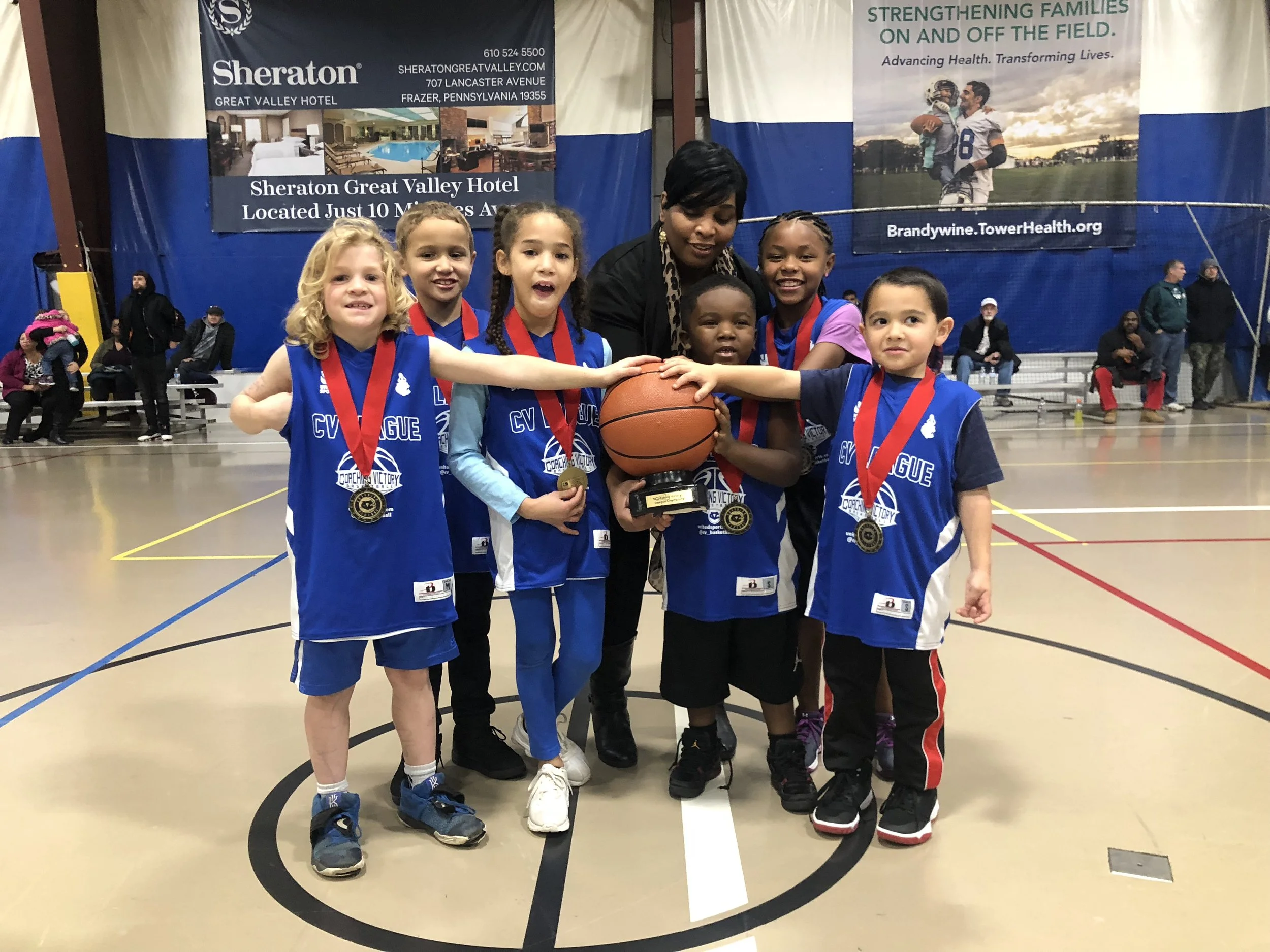 Young girls and their coach celebrate a basketball win, holding a trophy and wearing medals, inside a gymnasium with banners and spectators in the background.