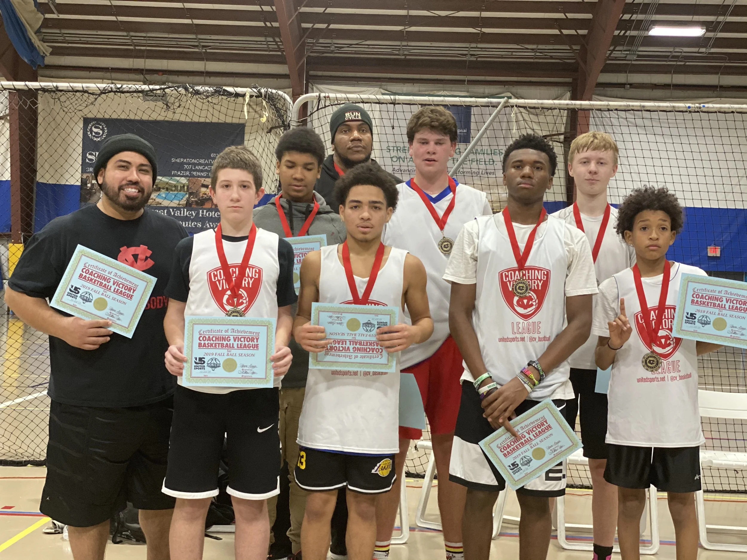 Group of young boys and a coach on an indoor basketball court, holding certificates and medals for winning a basketball league season.