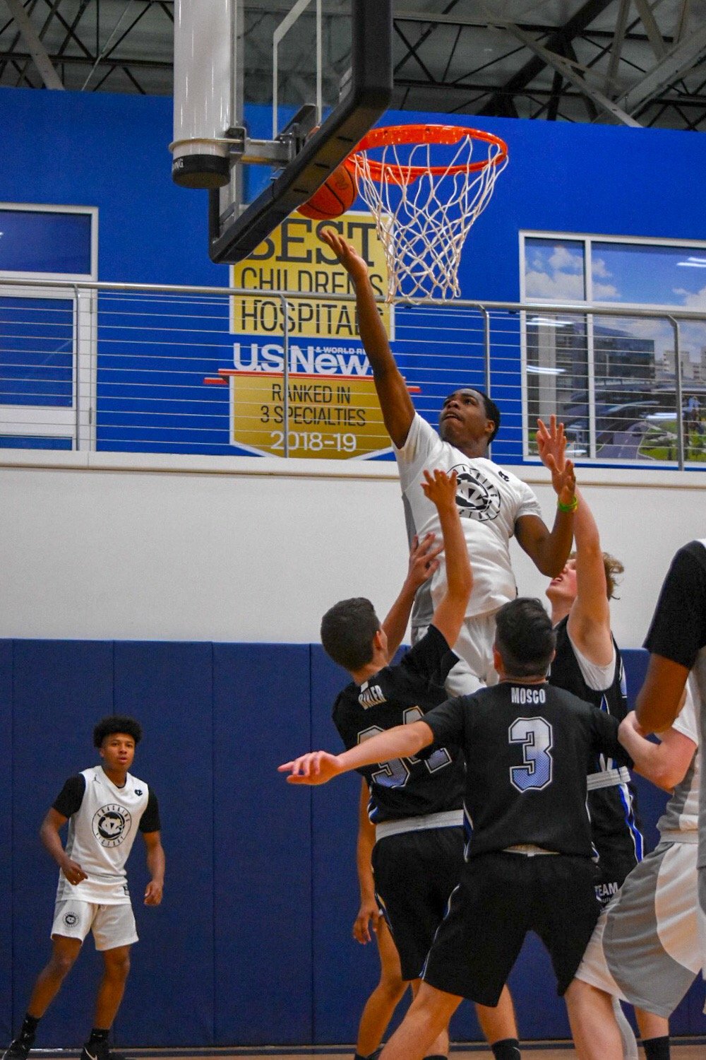 A group of boys playing basketball indoors, with one boy jumping to shoot the ball into the hoop. Others are attempting to block or defend, with a blue wall and a sign in the background.