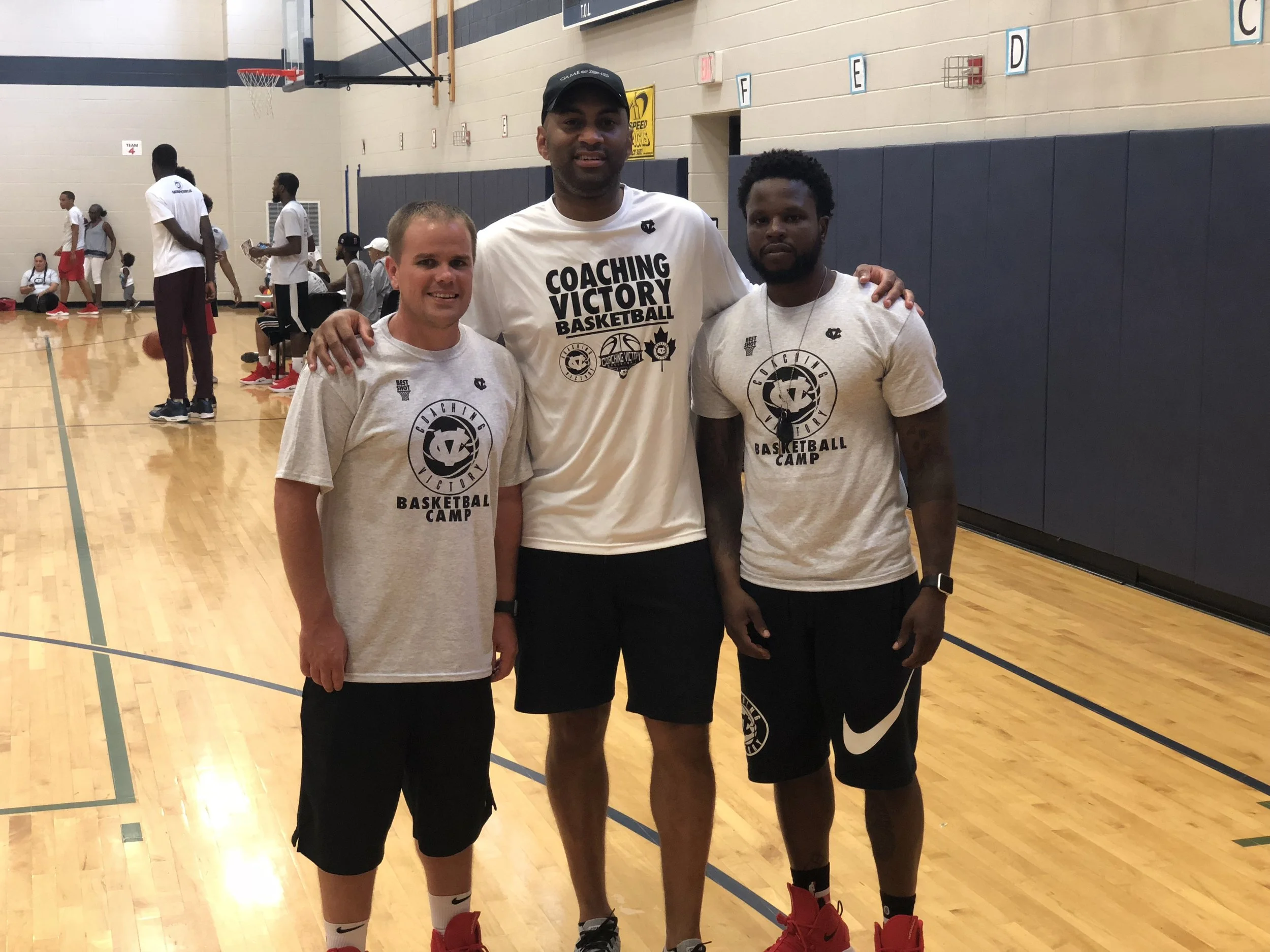 Three men standing together in a basketball gym, posing for a photo. The man in the center is tall and wearing a white shirt that says 'Coaching Victory Basketball.' The two men on each side are wearing grey 'Basketball Camp' t-shirts and black shorts.