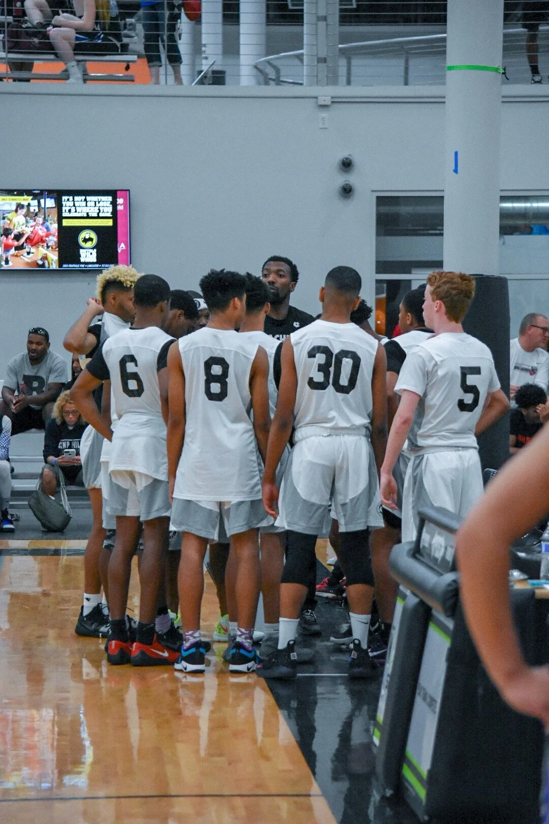 Basketball team in white uniforms listening to coach during timeout on indoor court.