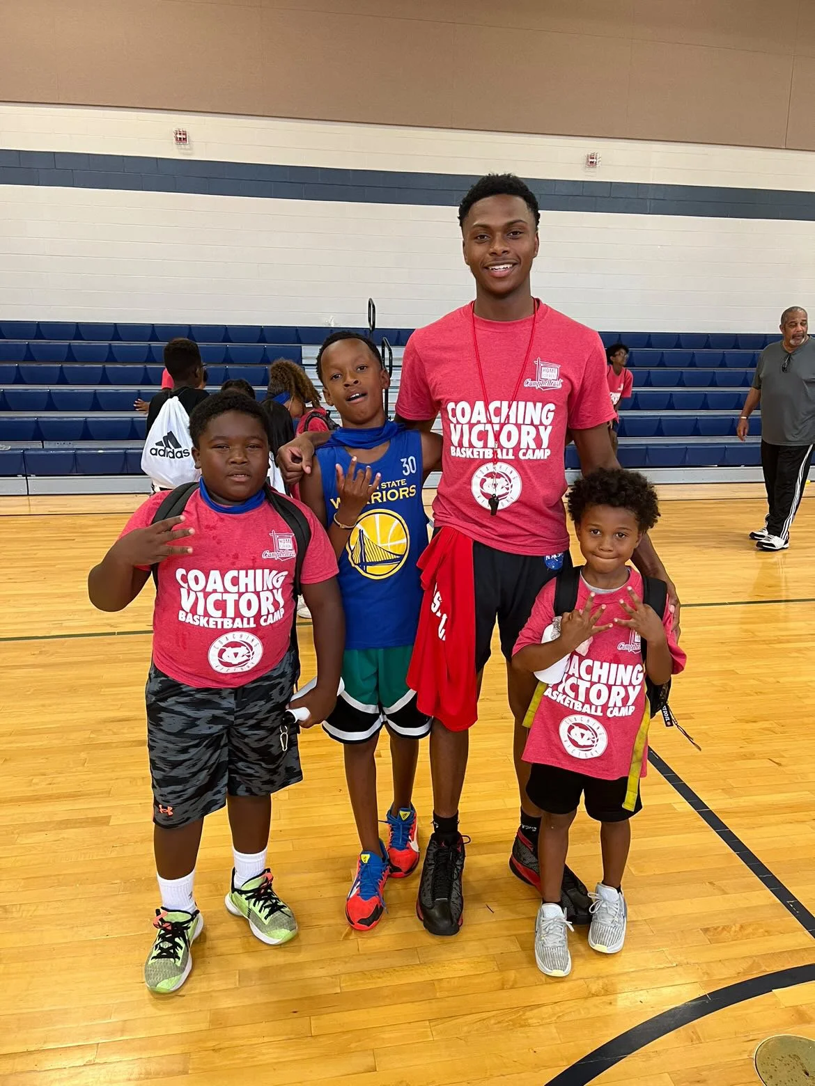 A group of four children and one adult male in a gymnasium, with bleachers in the background. The children are wearing sports attire, and two of them have red shirts that say 'Coaching Victory Basketball Camp.' The adult male is standing behind them, smiling.