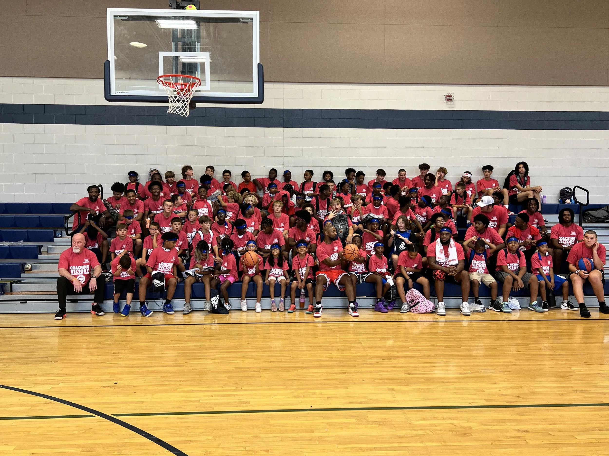 Group of children and adults sitting on bleachers in a gymnasium, wearing matching pink t-shirts with 'Coaching Victory' written on them, with a basketball hoop overhead.
