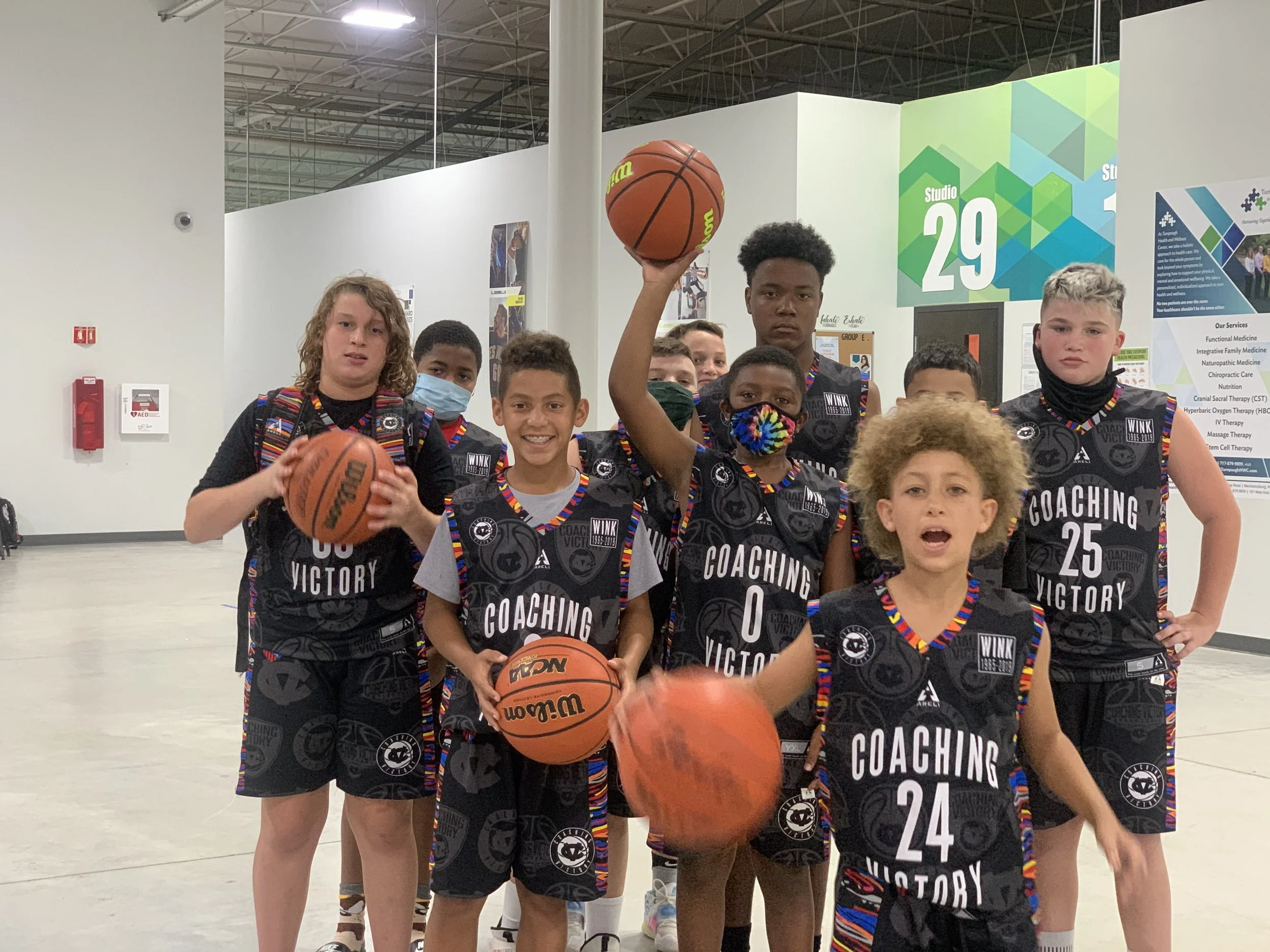 Group of young children wearing black basketball jerseys with colorful trim, holding basketballs, inside a sports arena or gym.