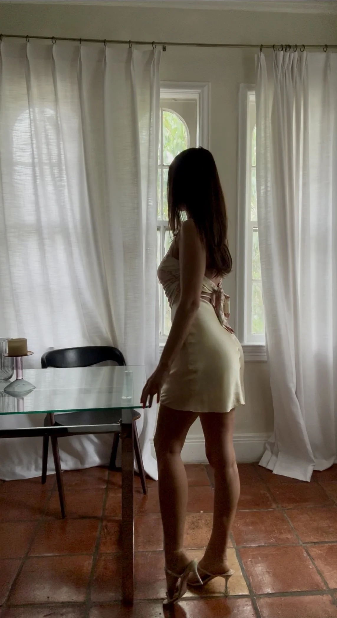 A woman in a beige dress and high heels standing in a room with white curtains and a glass table.