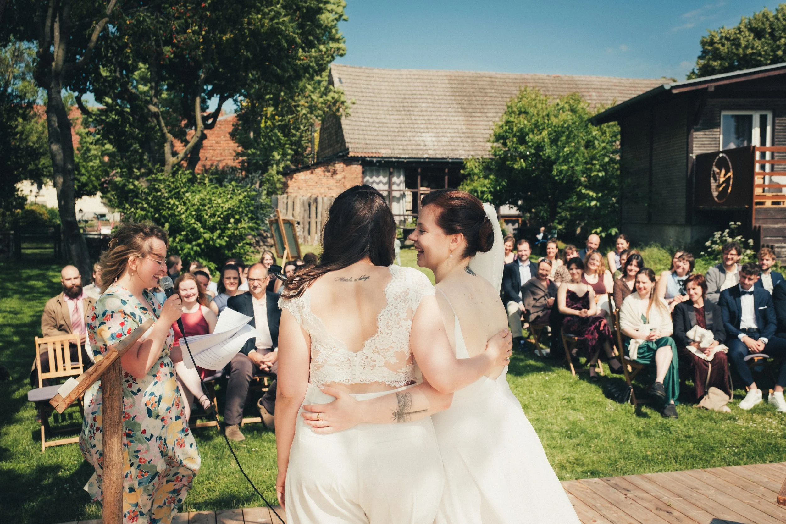 Two brides kissing at an outdoor wedding ceremony with guests seated on chairs behind them, trees, and a rustic building in the background.