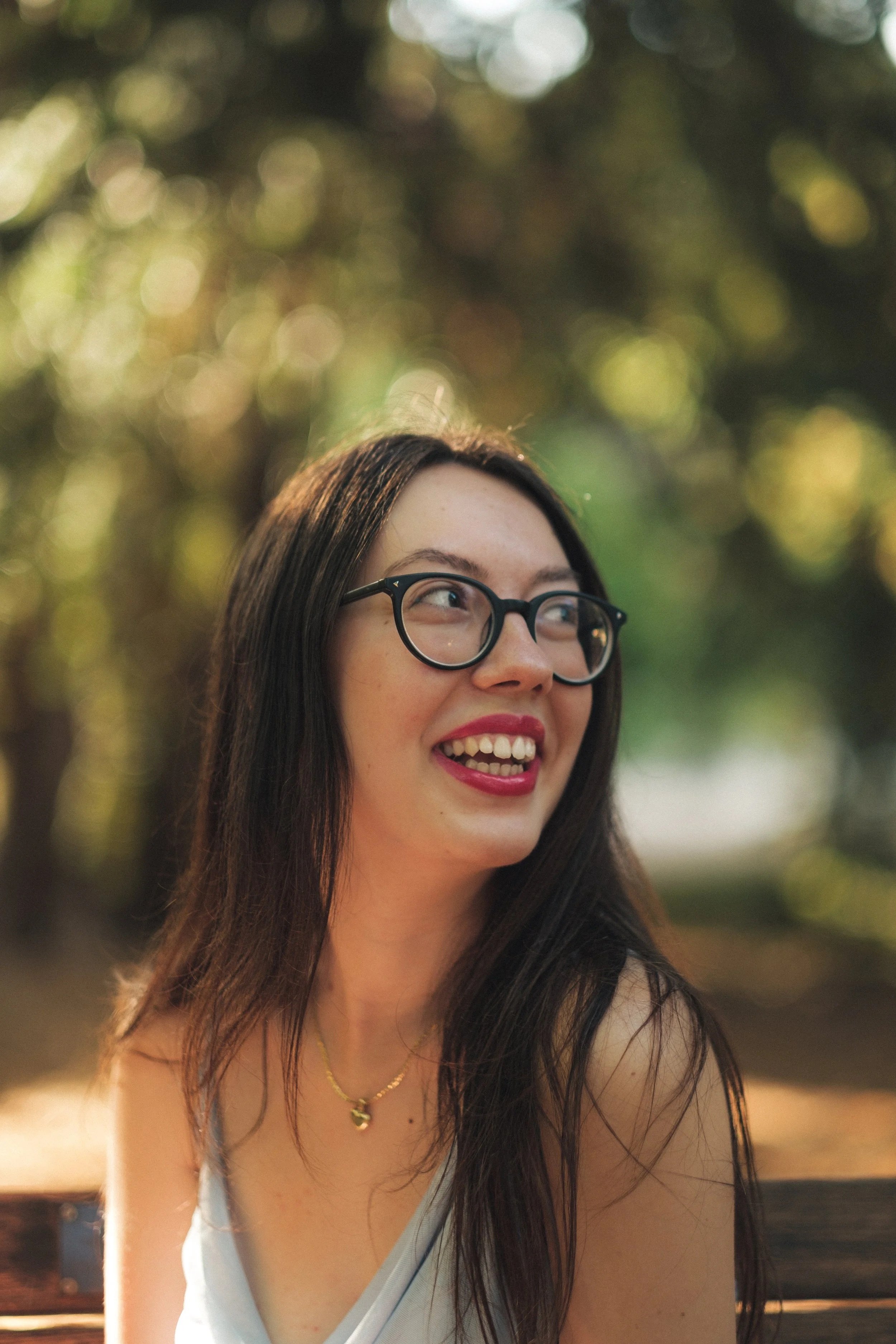 A woman with glasses and red lipstick smiling outdoors, with blurred green and gold foliage in the background.