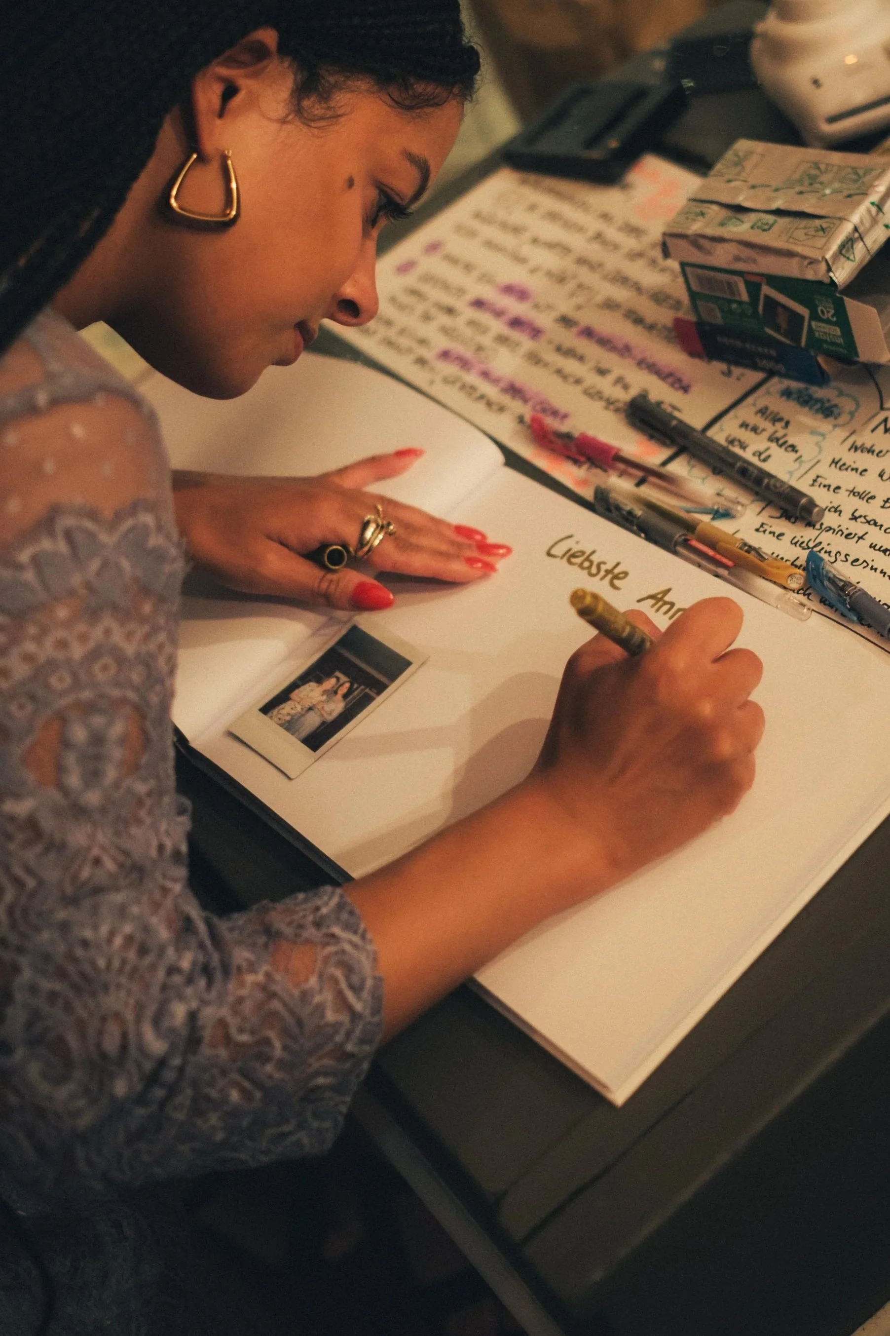 A woman with dark hair, gold earrings, and red nail polish, writing a message on a white paper that reads 'Liebste An...' surrounded by pens, tissue paper, and a Polaroid photo on a table.