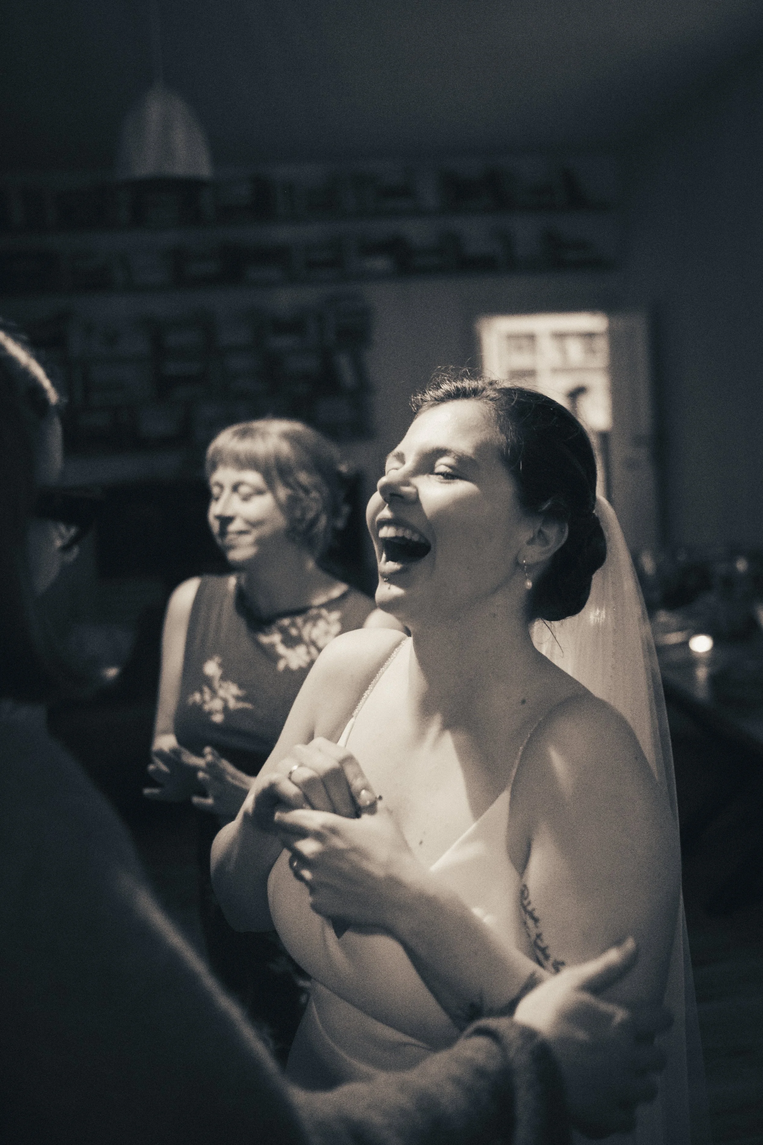 Black and white photo of a woman with dark hair styled in an elegant updo, laughing and wearing a sleeveless dress. She is standing next to another woman with short hair and a floral dress, in a room with a window and exposed brick wall.
