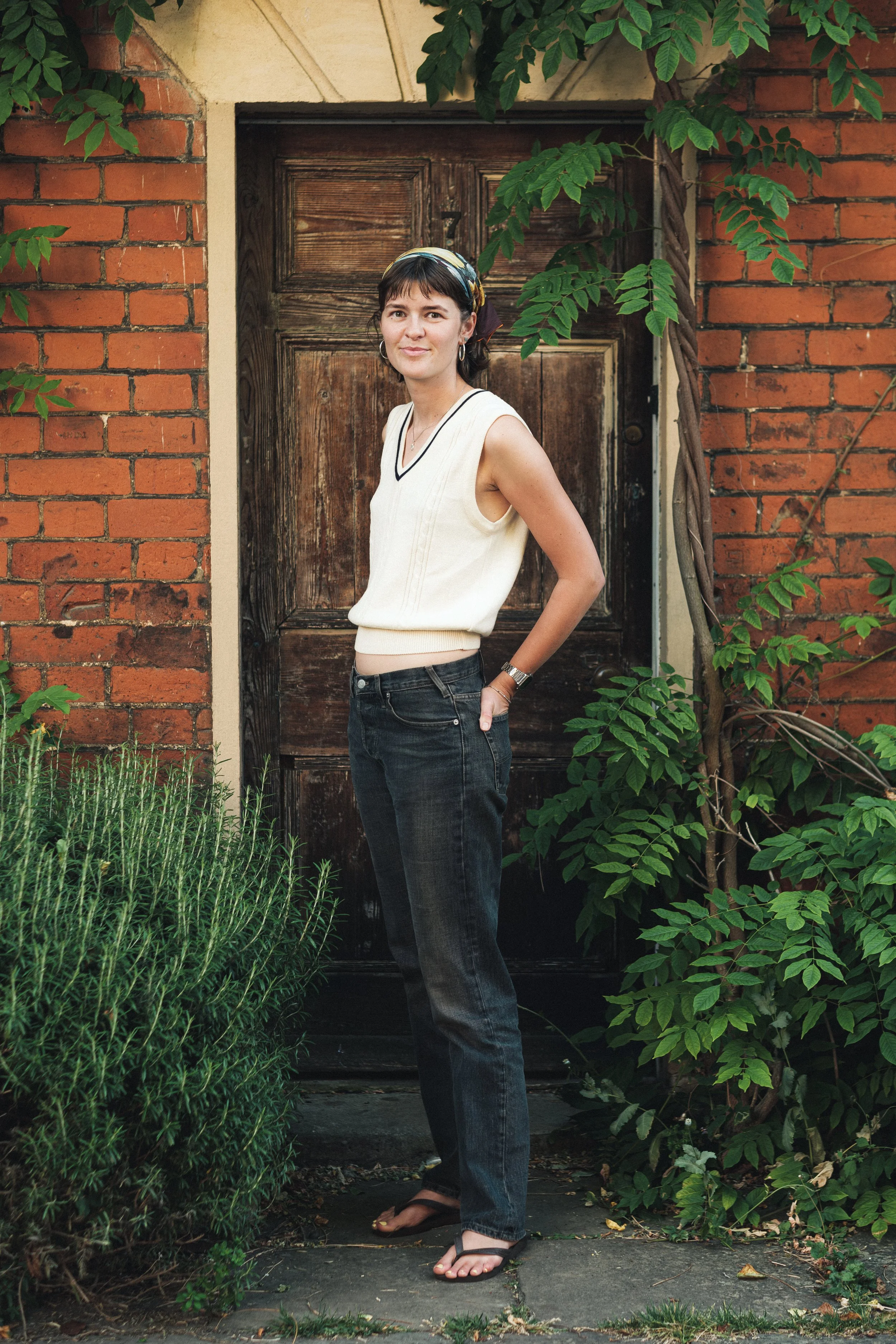 A woman standing on a stone pathway in front of a wooden door, surrounded by green foliage and a brick wall.
