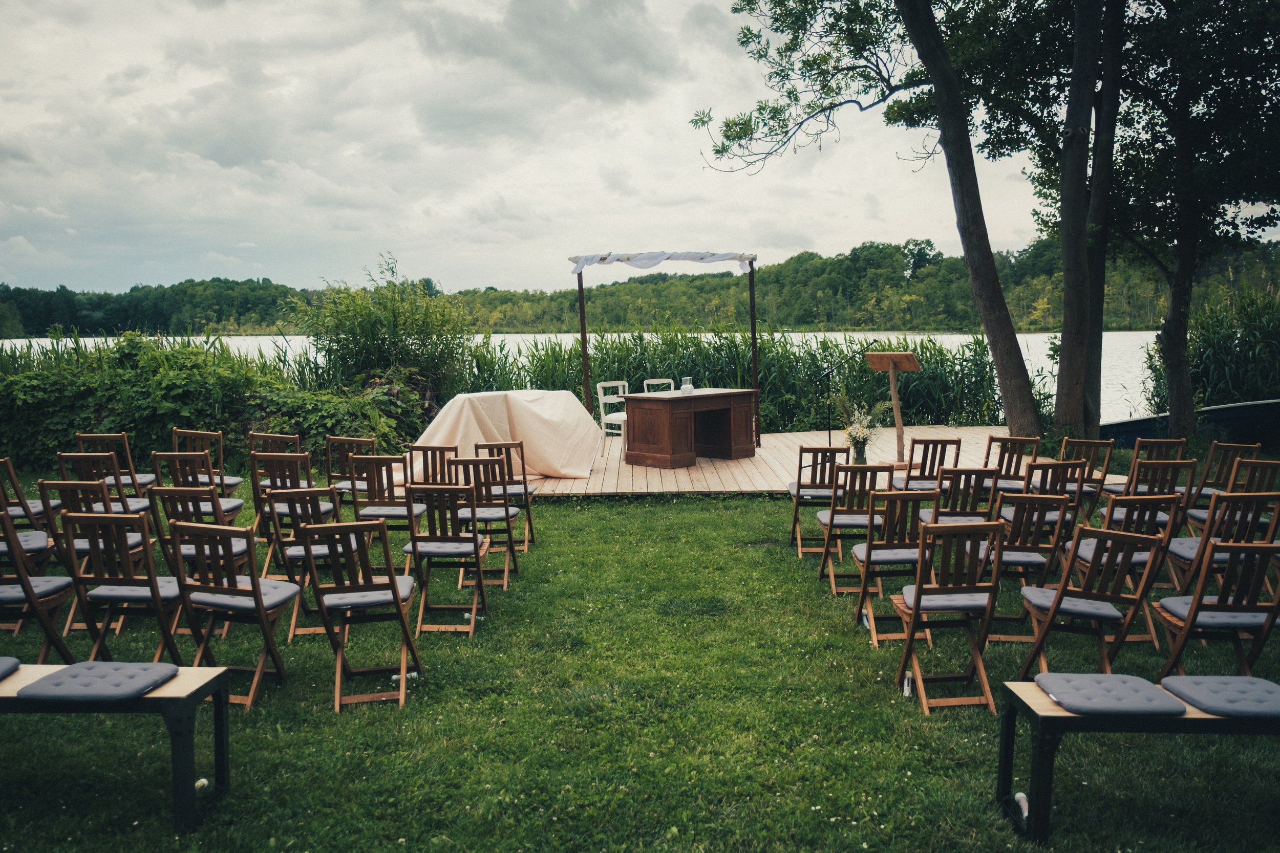 Outdoor wedding setup with chairs arranged on grass facing a small wooden platform with a canopy, overlooking a lake with trees and cloudy sky in the background.
