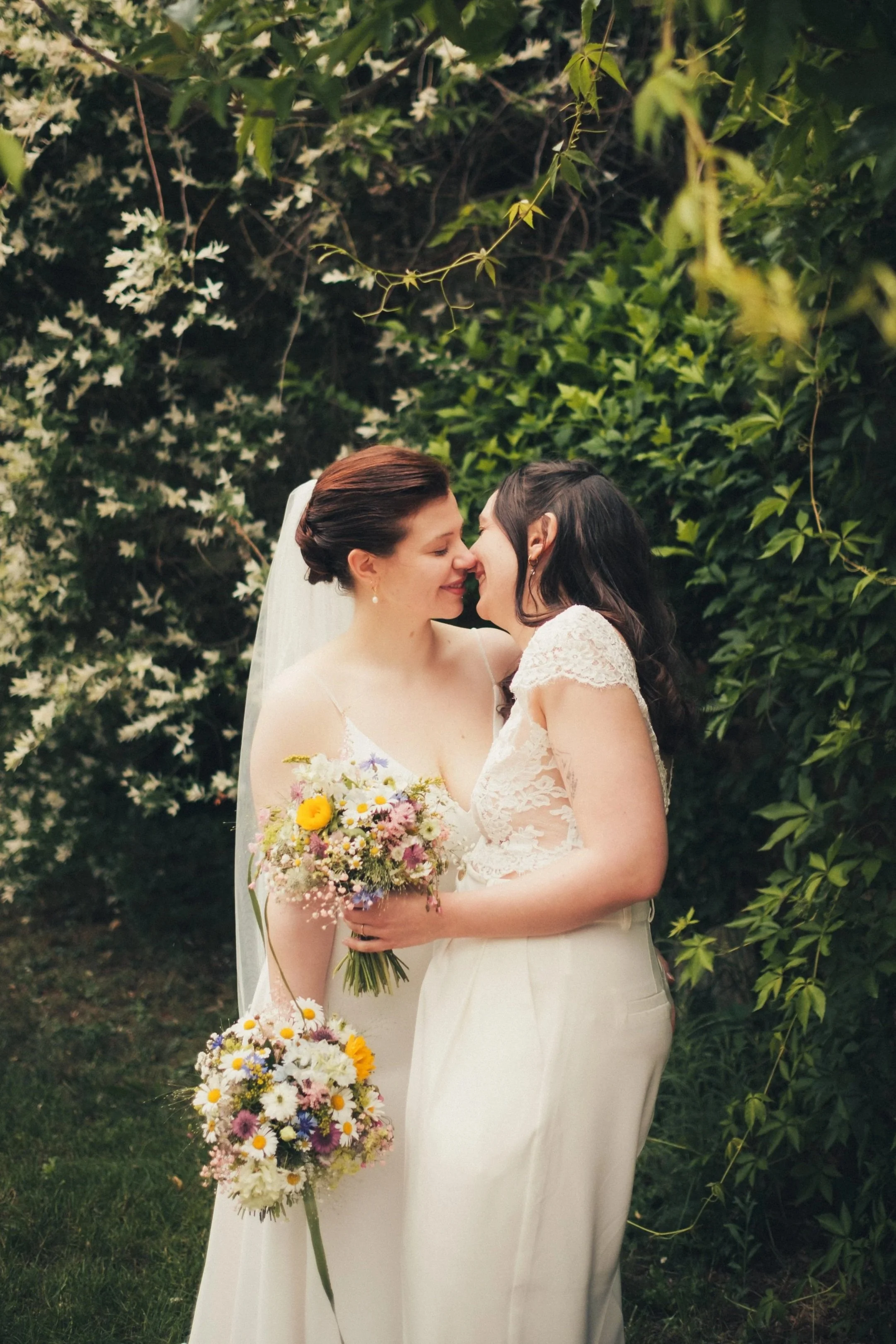 Two women in wedding dresses smiling and touching noses outdoors, surrounded by green foliage, with one woman holding a bouquet of colorful flowers.