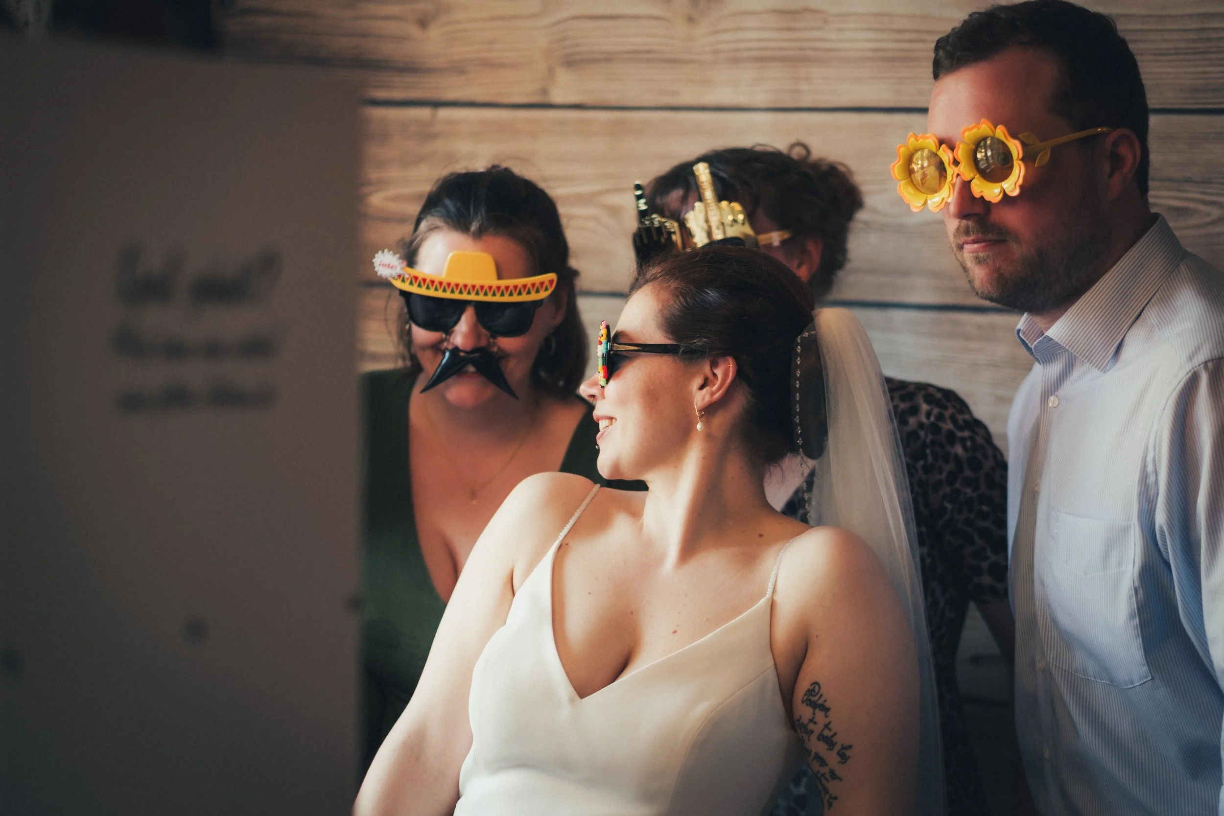 A group of people at a party wearing playful sunglasses and costumes, with one person wearing a fake mustache and sombrero sunglasses, and a woman with a dress and sunglasses enjoying the event