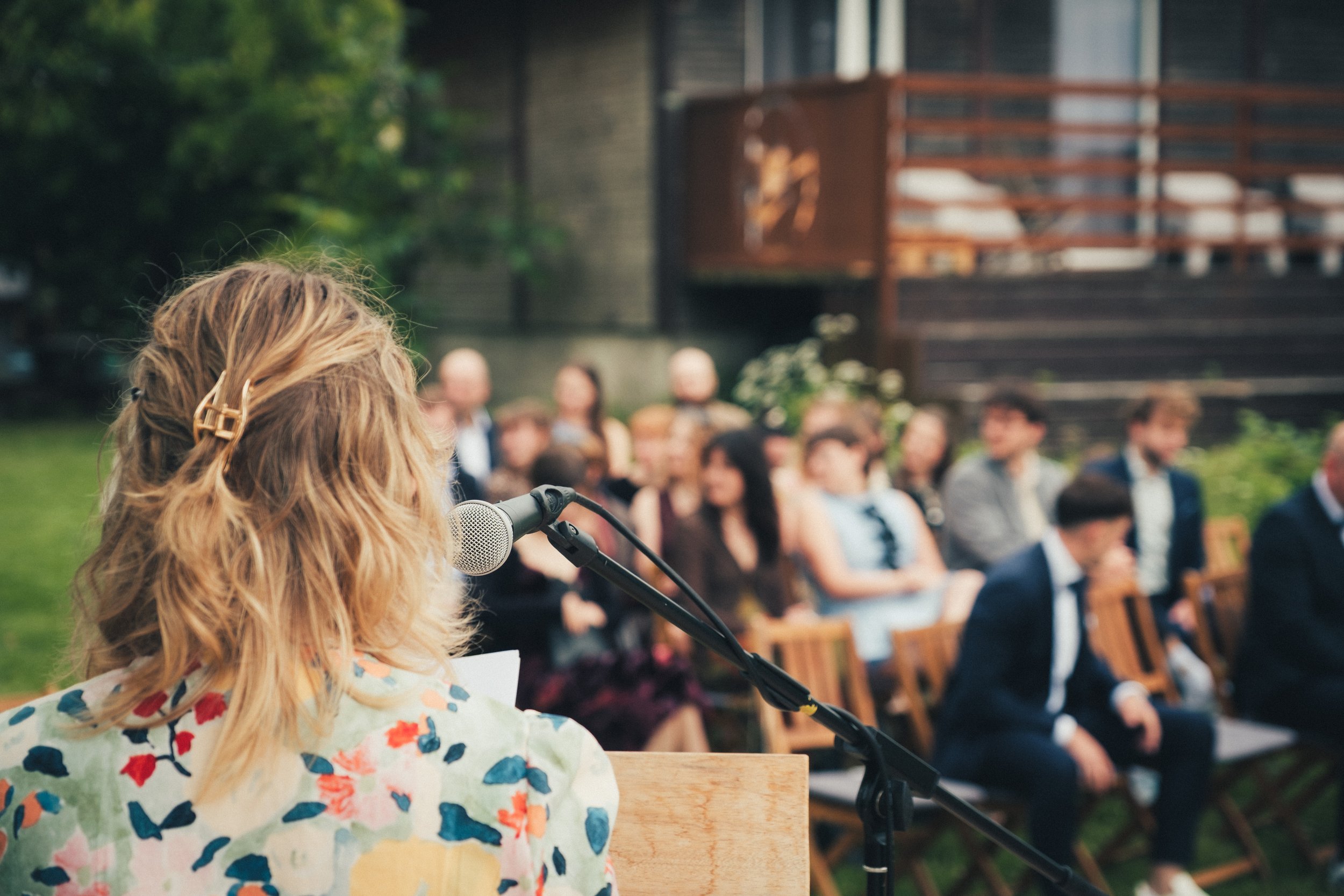 A woman with wavy blonde hair, wearing a floral dress, stands at a microphone during an outdoor event. In the background, a group of people is seated on chairs, and there is a wooden deck and greenery.