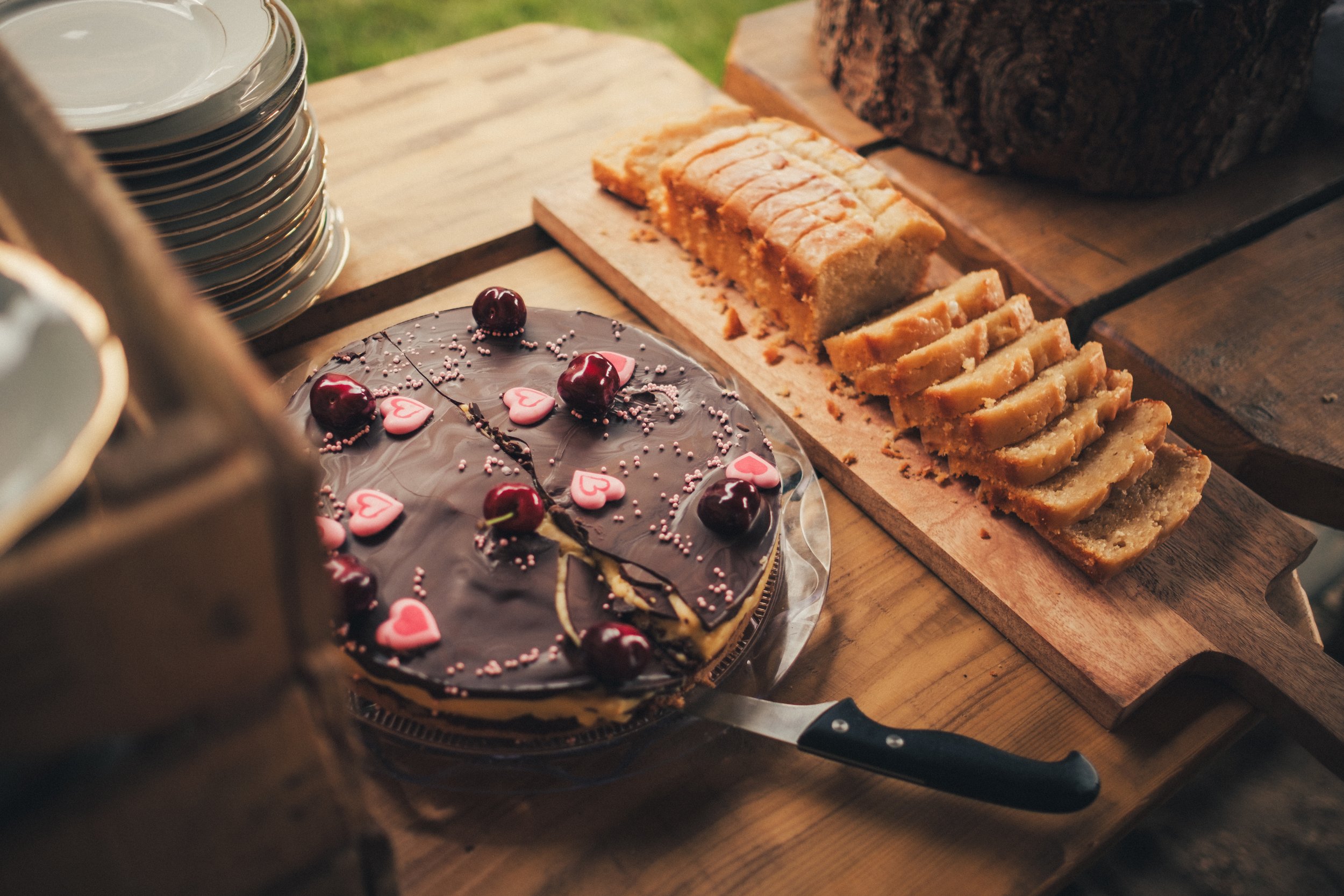Chocolate cake with cherries and pink heart-shaped decorations on top, placed on a clear cake stand. On a wooden cutting board, there are sliced pound cakes and a loaf of bread. A stack of white bowls is visible on the left side.