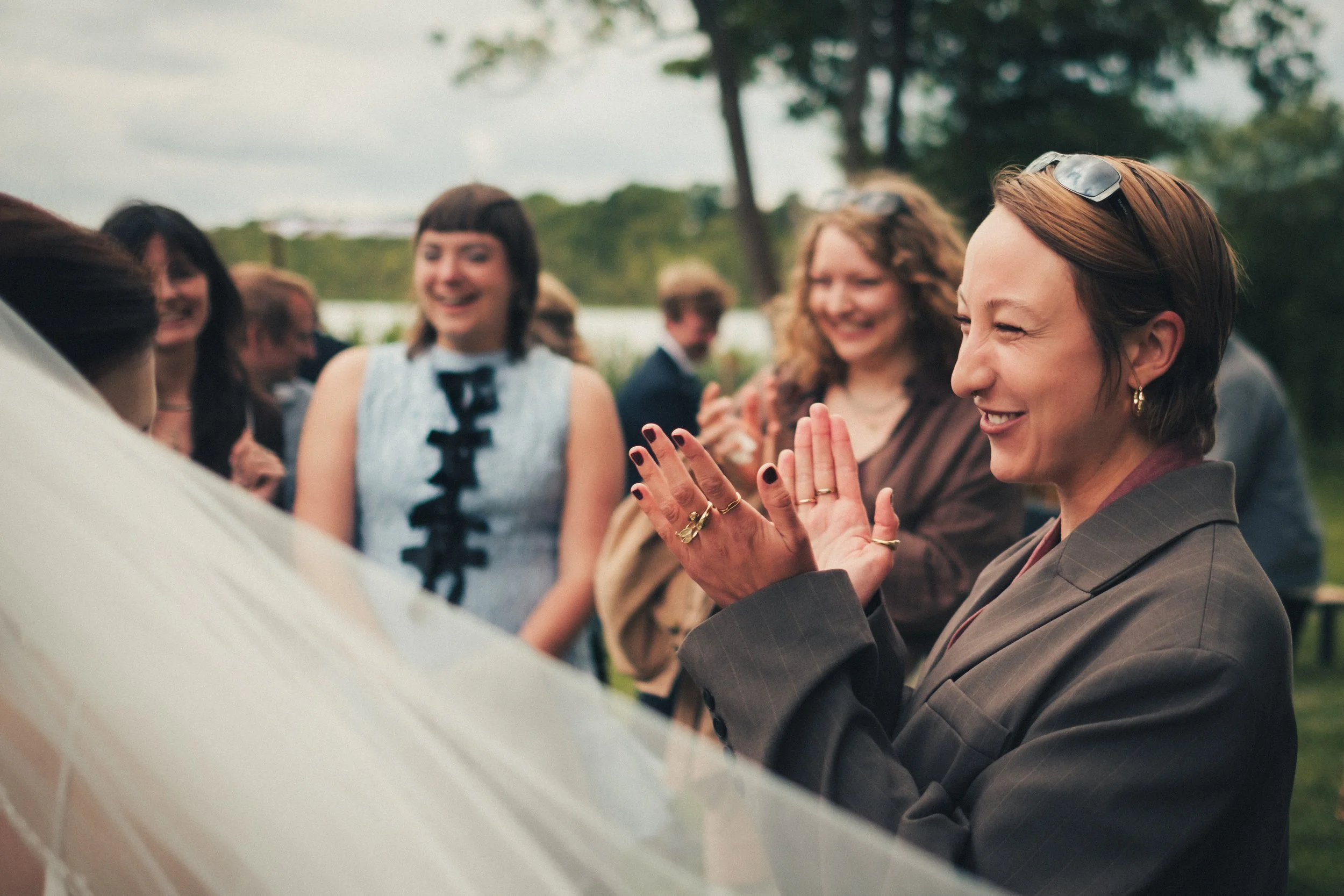 A group of women outdoor, with one woman in a gray blazer clapping. Others are smiling and talking, with trees in the background.