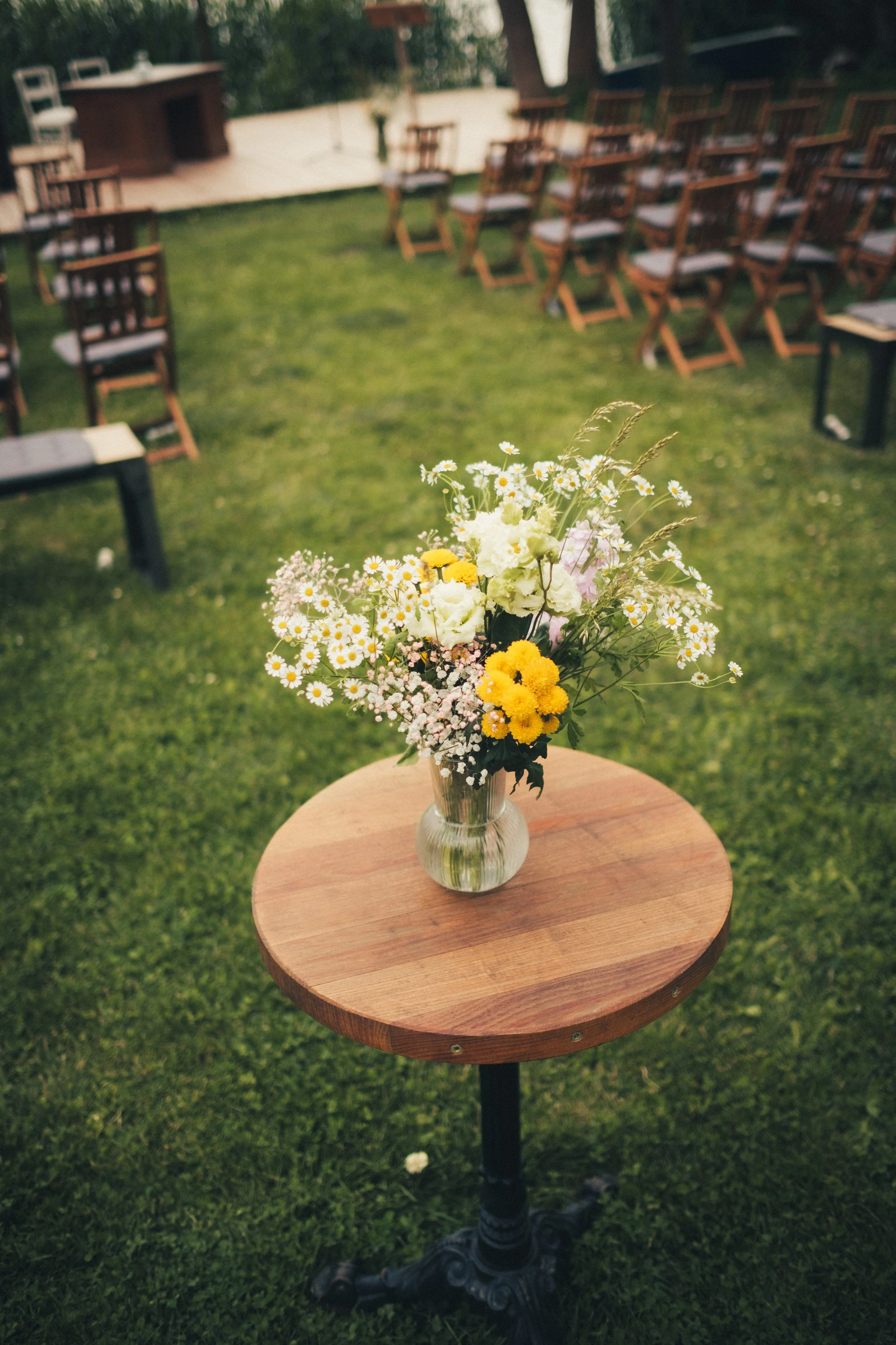 A small wooden round table with a vase of mixed wildflowers on top, located outdoors on a grassy area.