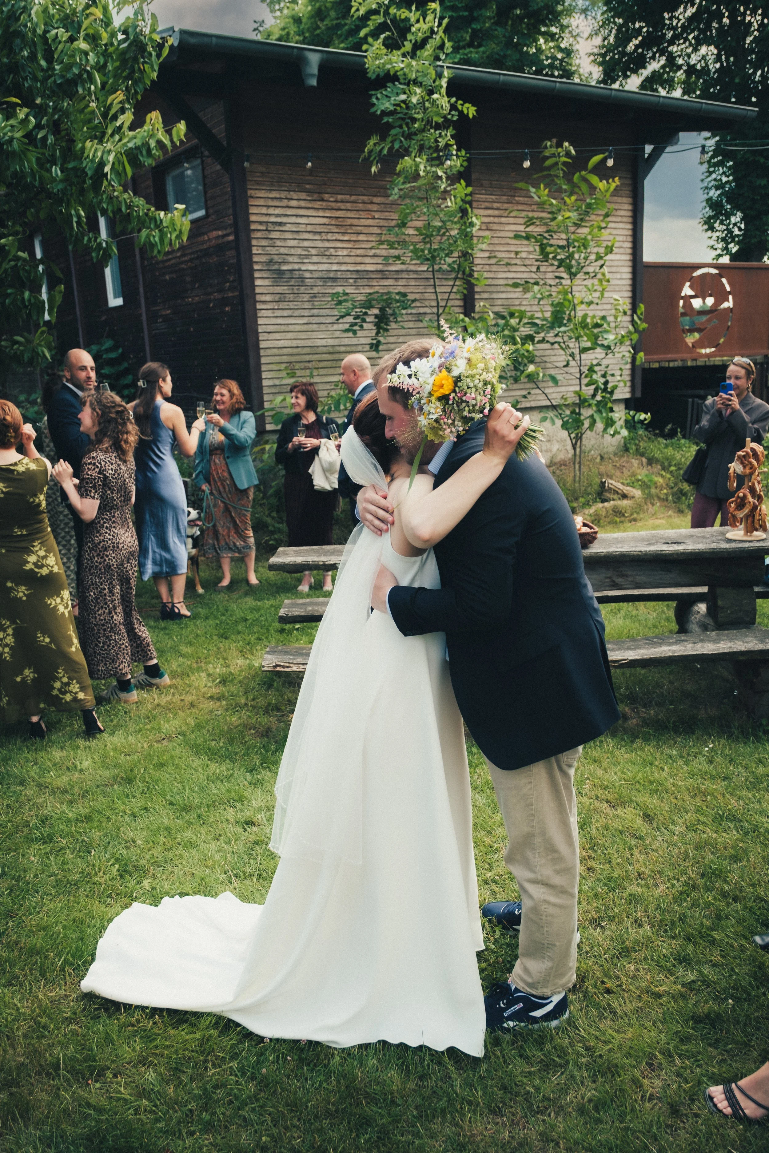 A wedding celebration outdoors with the bride and groom hugging, surrounded by guests. The bride is holding a colorful bouquet, and the groom is wearing a dark blazer. Guests are talking and taking photos in a green grassy area with a wooden building in the background.