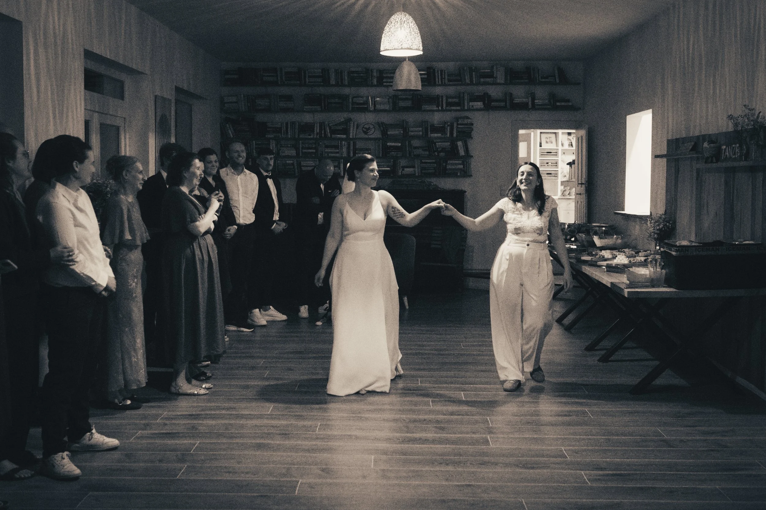 A group of people dressed in formal attire standing in a room, with two women in the center holding hands and smiling, likely celebrating a special occasion.