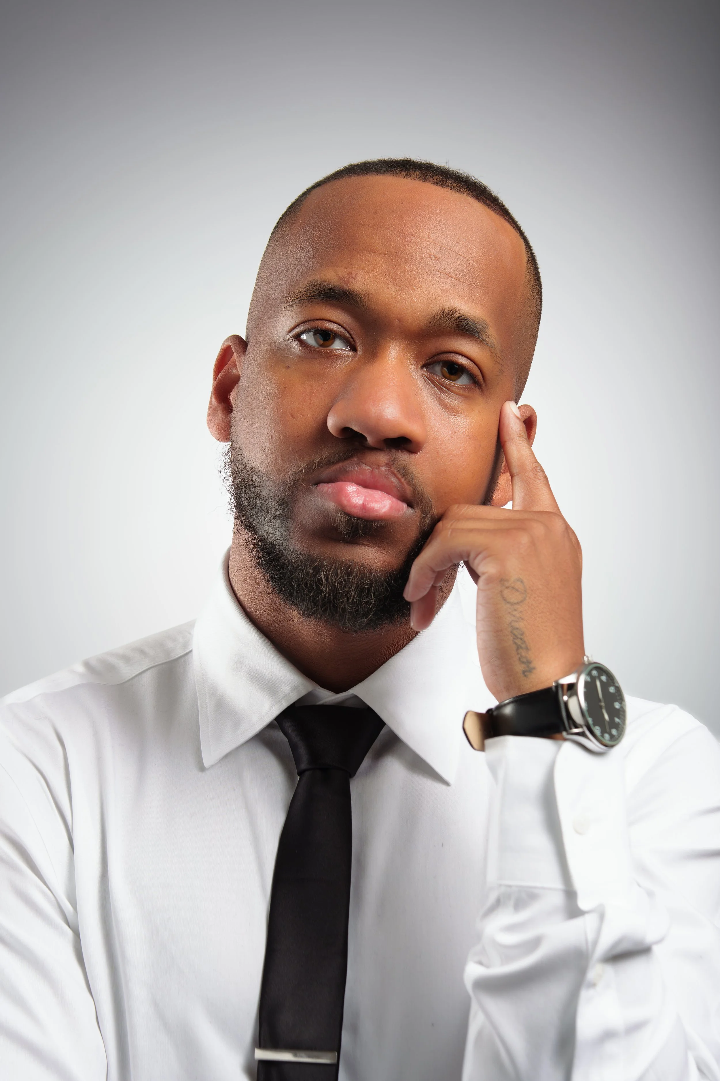 A man in a white shirt and black tie with a watch, resting his head on his hand, looking at the camera with a serious expression.