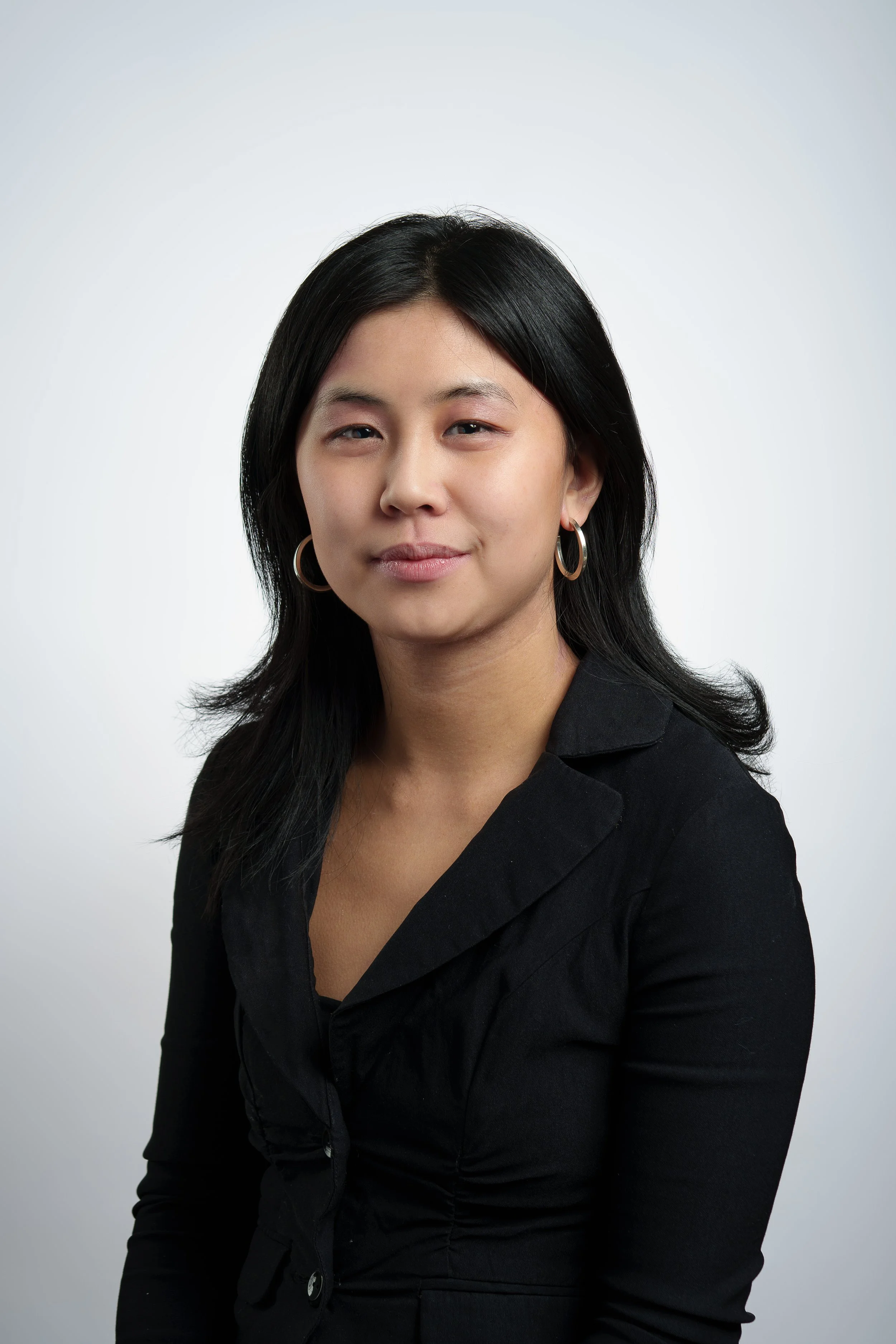 A woman with black hair, wearing hoop earrings and a black blazer, posing against a plain light-colored background.