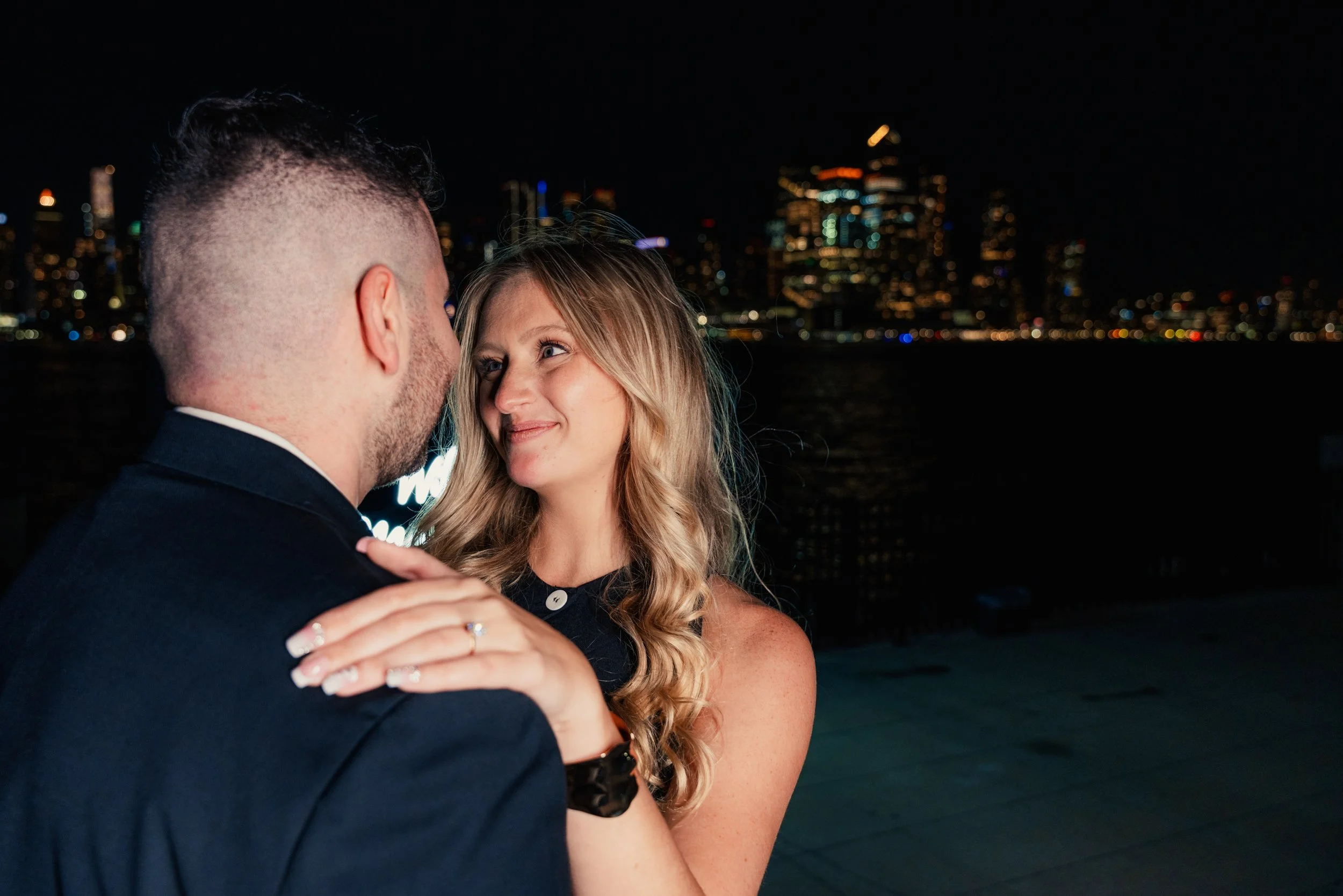 A couple dancing closely on a rooftop at night with city lights in the background.