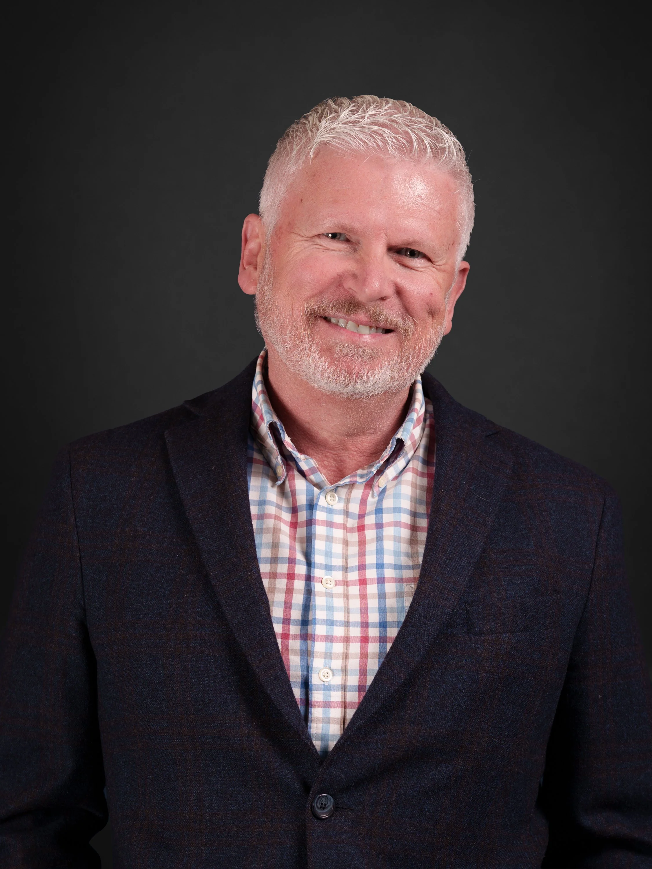 A smiling older man with white hair and beard wearing a dark blazer and a checkered shirt, posing in front of a dark background.