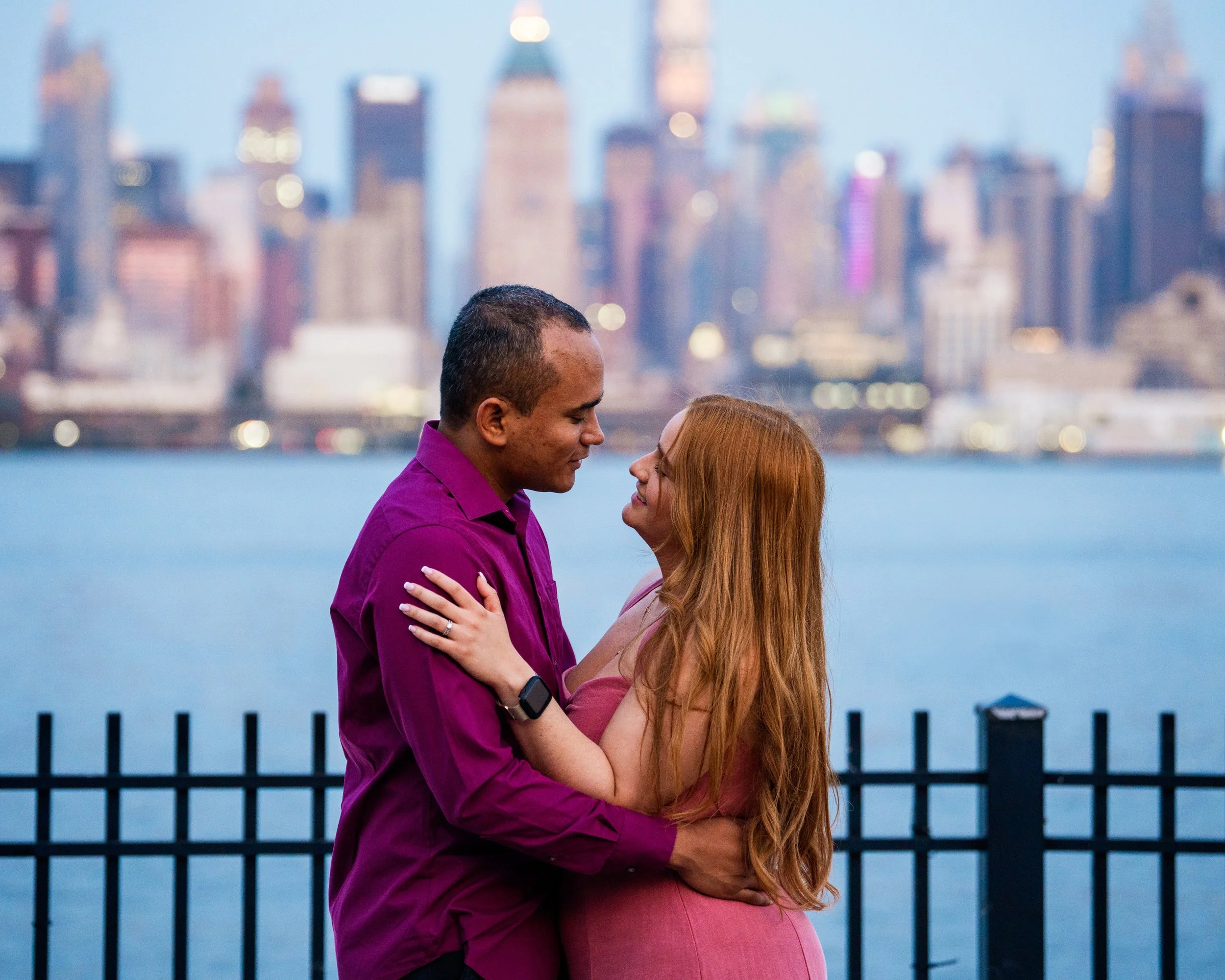 A couple embracing by a waterfront with a city skyline in the background.