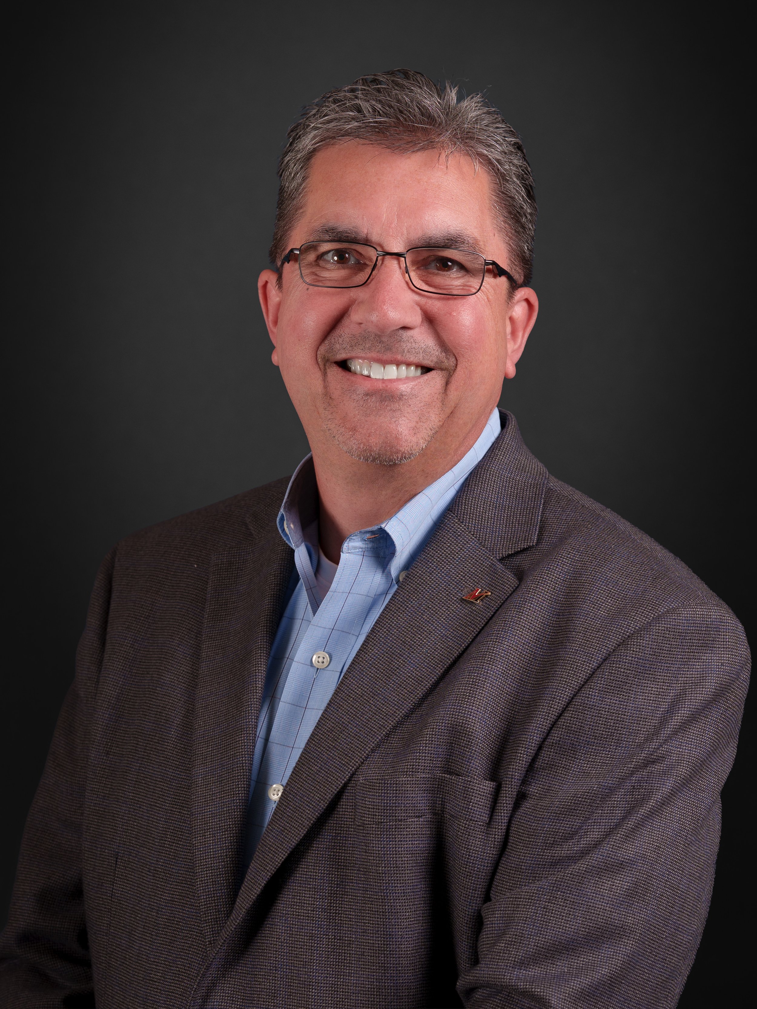 A professional headshot of a middle-aged man with glasses, smiling, wearing a brown blazer over a light blue shirt, against a dark background.