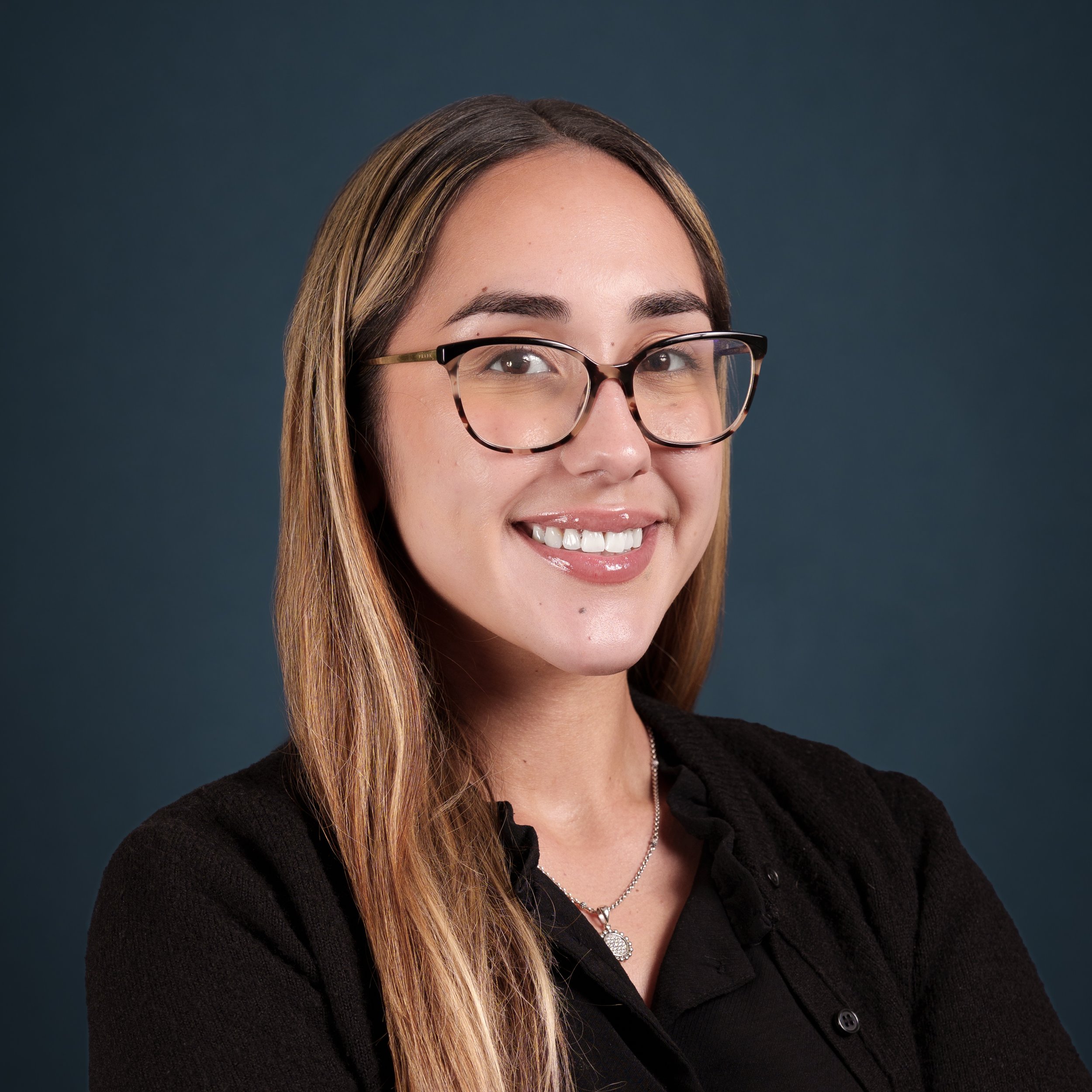 Portrait of a young woman with light skin, long wavy hair, glasses, and a black top, smiling against a dark teal background.