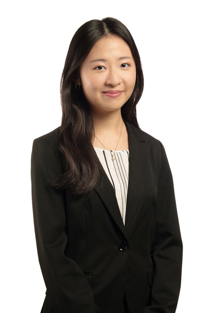 A young woman with long dark hair, wearing a black blazer over a white shirt with black vertical stripes, standing against a white background.