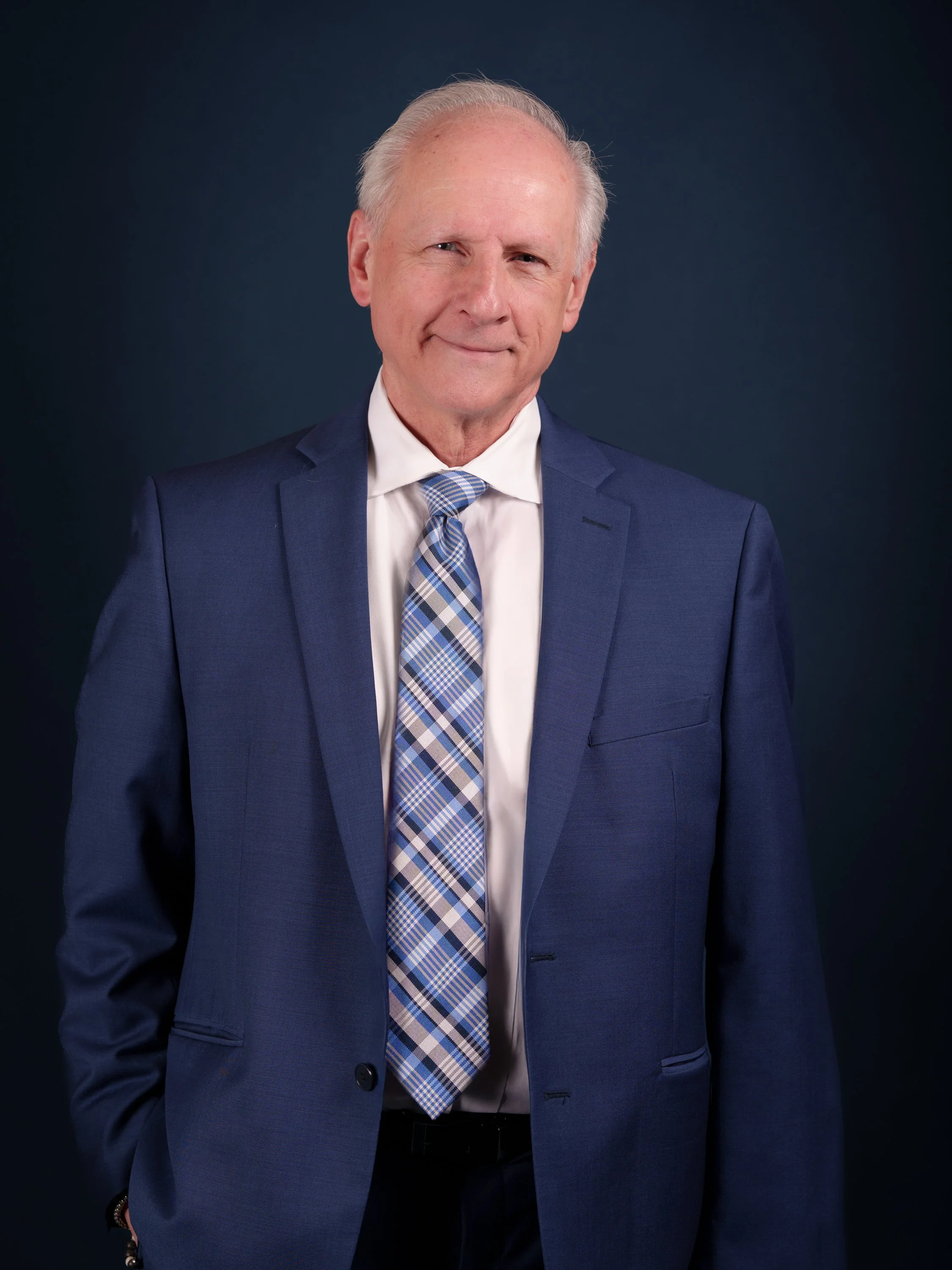 An older man with gray hair wearing a dark blue suit, white shirt, and a plaid blue and white tie, standing against a dark background.