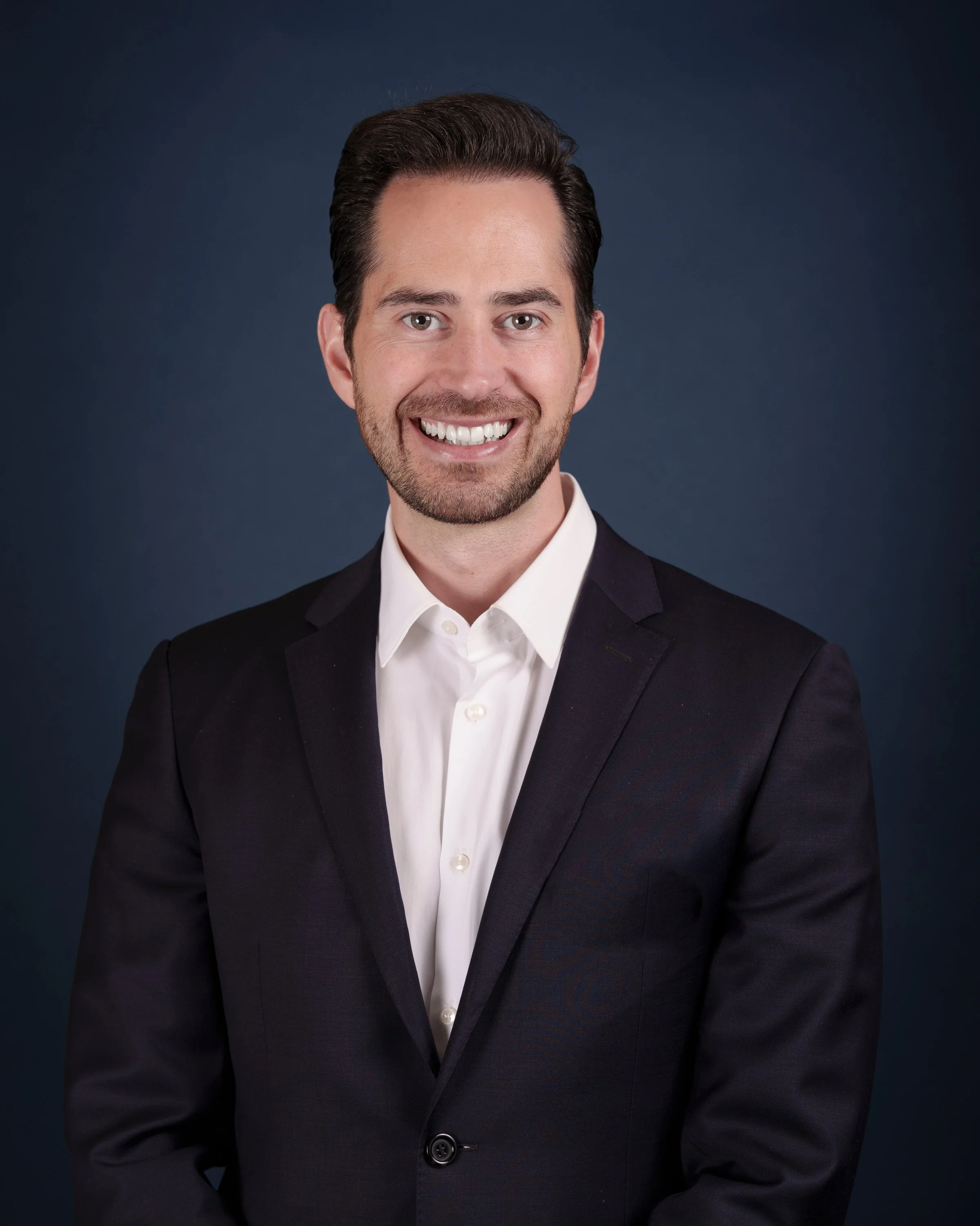 A portrait of a man with dark hair and a beard, smiling, wearing a black suit jacket and a white dress shirt, against a dark blue background.