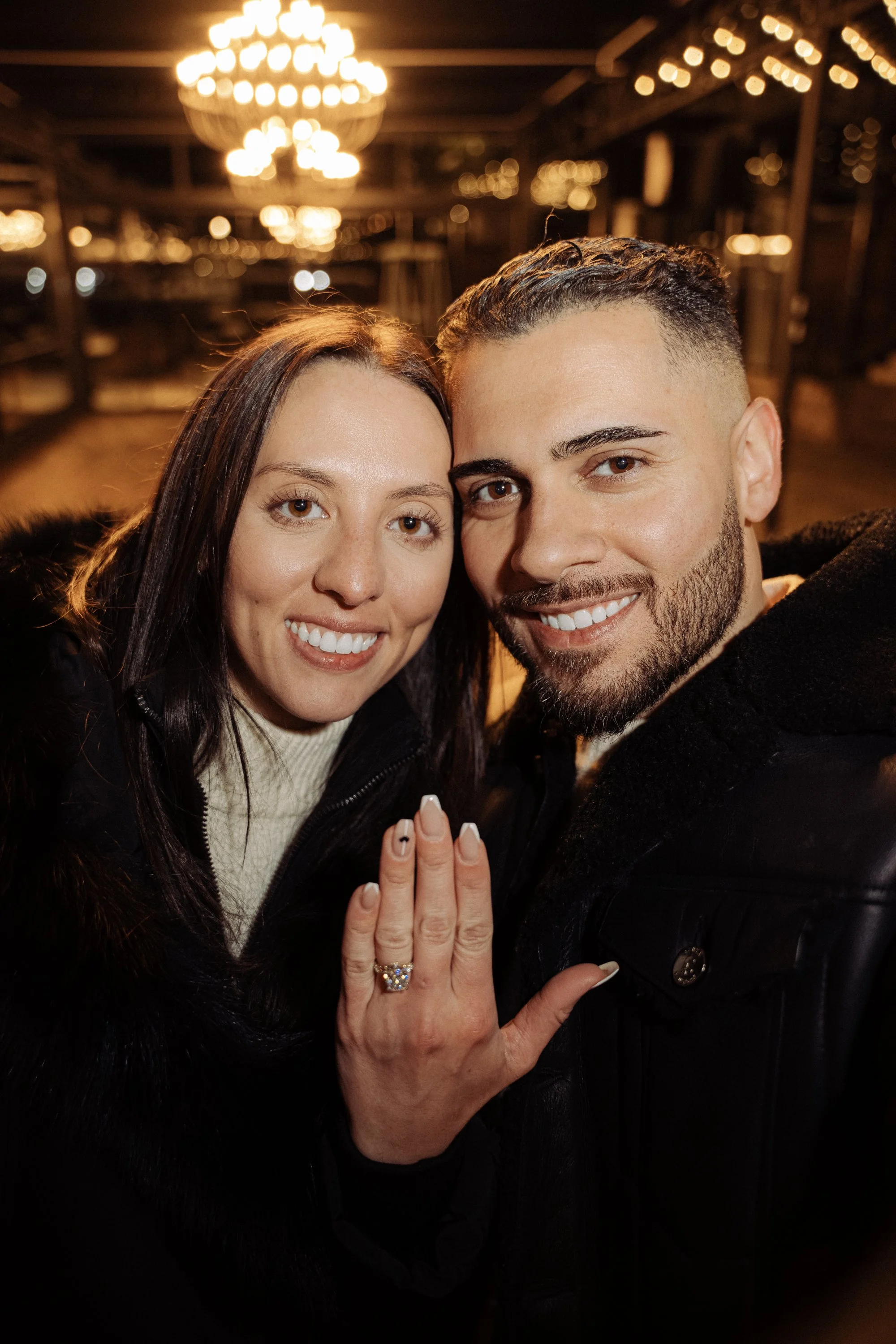 A smiling couple taking a selfie at night, with the woman's left hand showing an engagement ring.