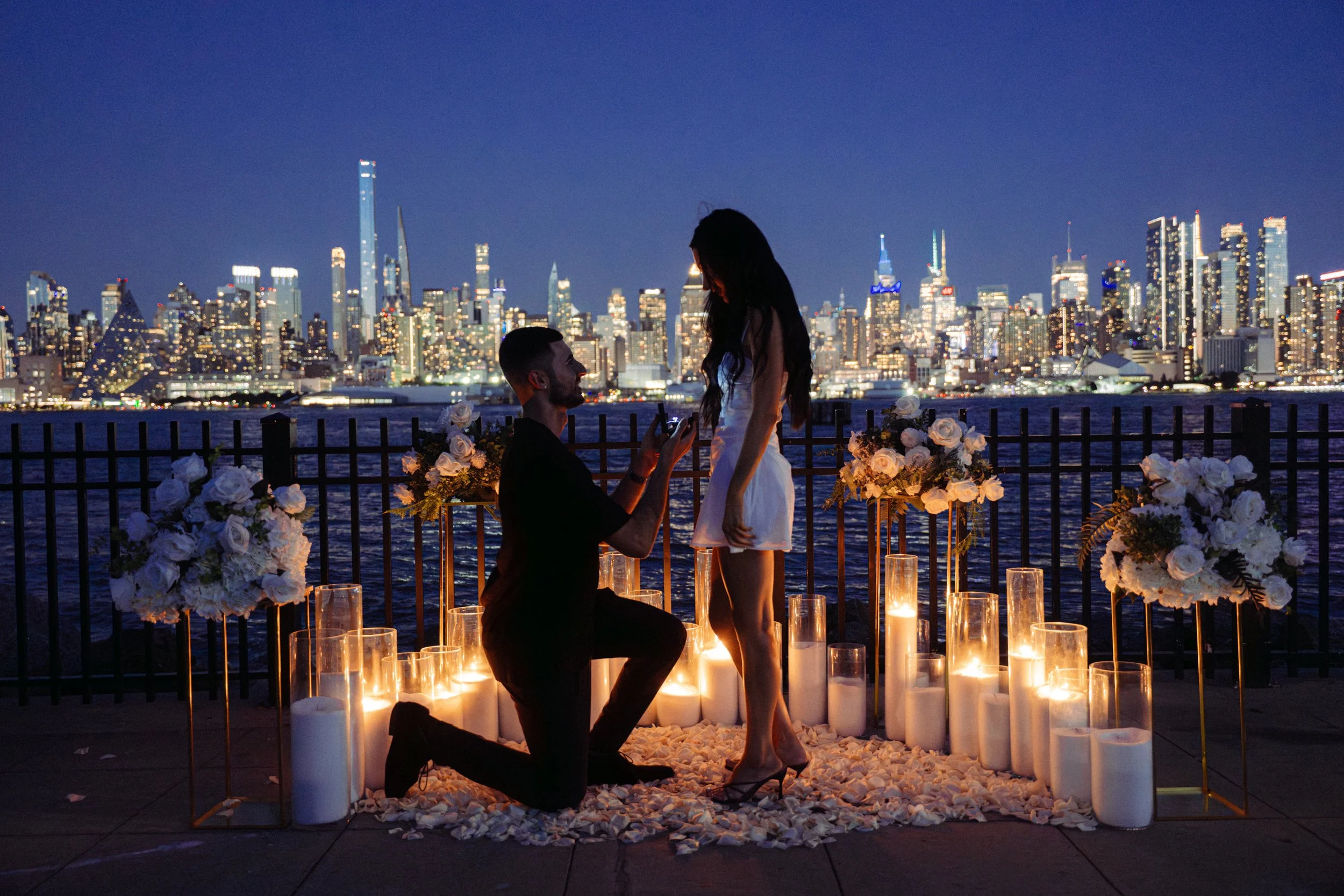 A couple proposes on a rooftop with a city skyline at night, surrounded by candles and floral arrangements.