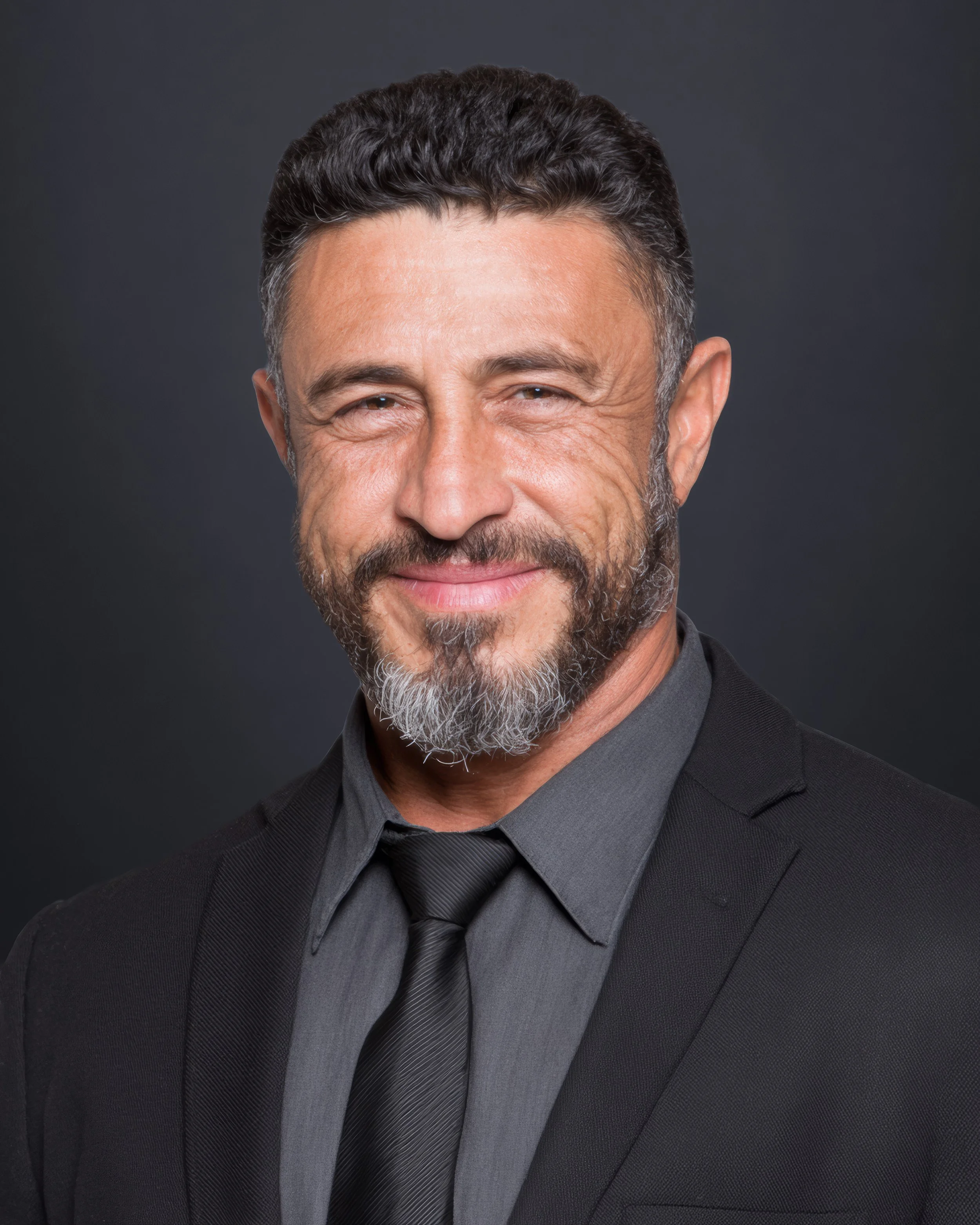 A smiling middle-aged man with a beard and short dark hair, dressed in a black suit, gray shirt, and black tie, posing against a dark background.