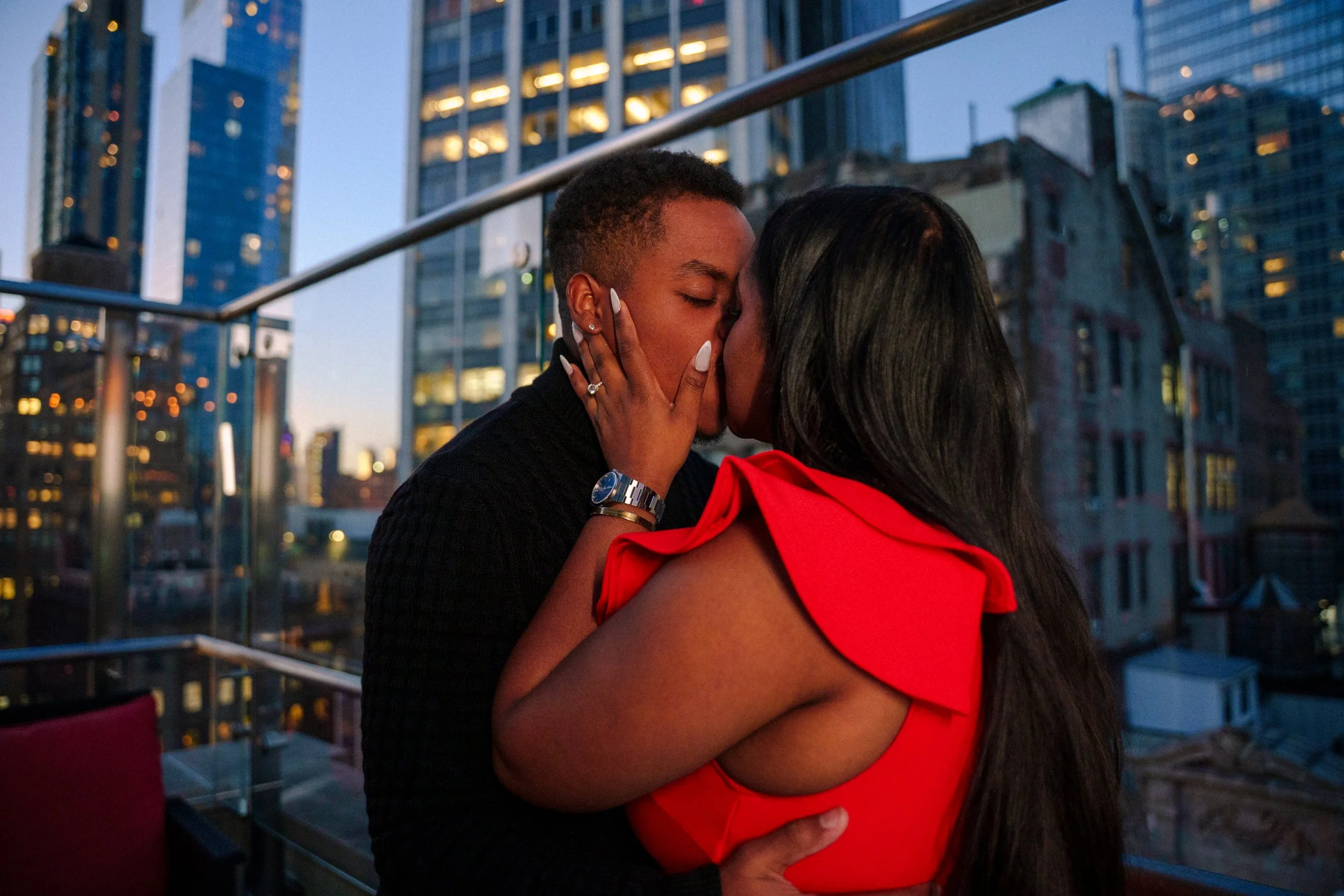 A couple kissing on a rooftop at dusk with city skyscrapers in the background.