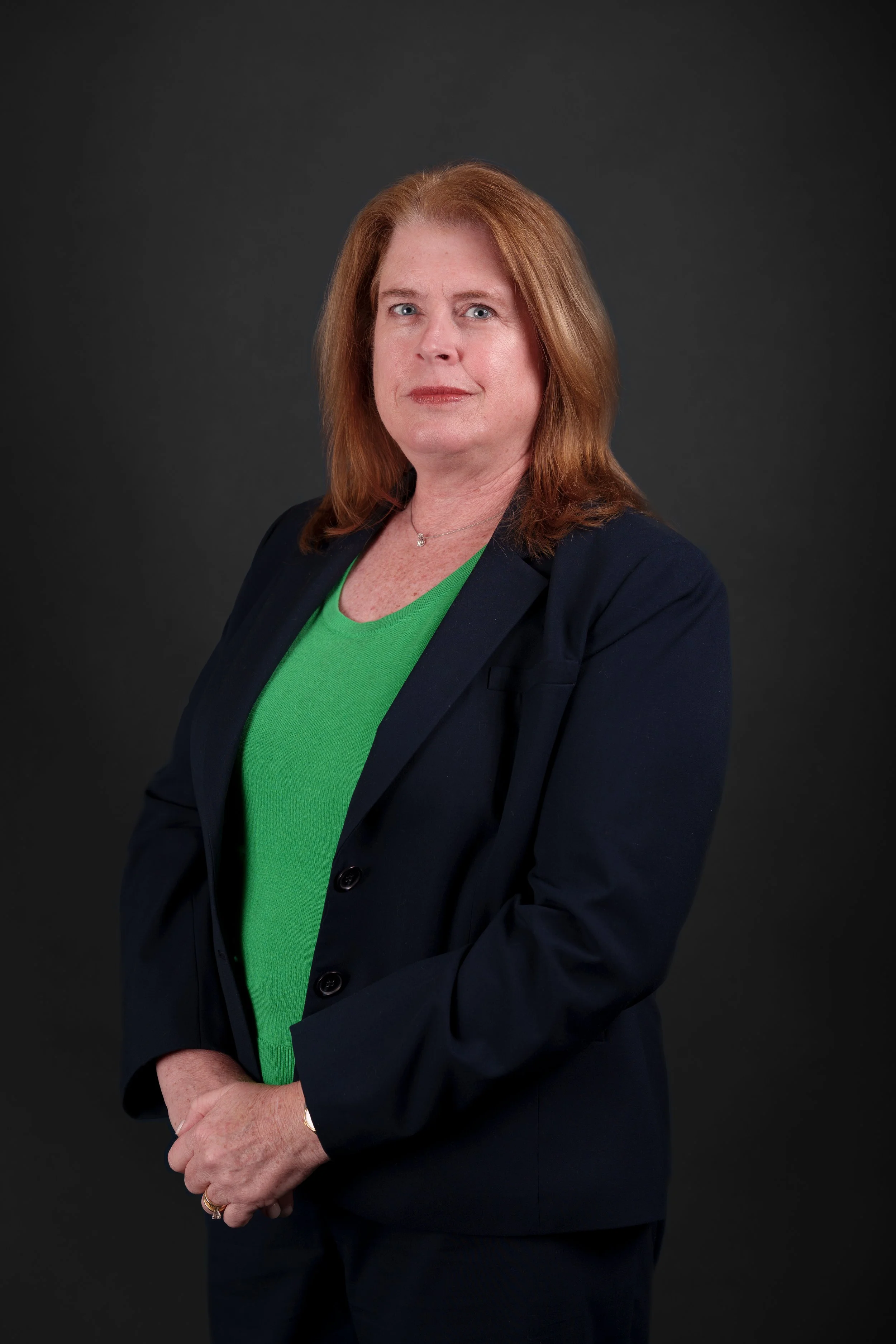 A professional woman with red hair, wearing a black blazer and a green top, standing against a dark background.