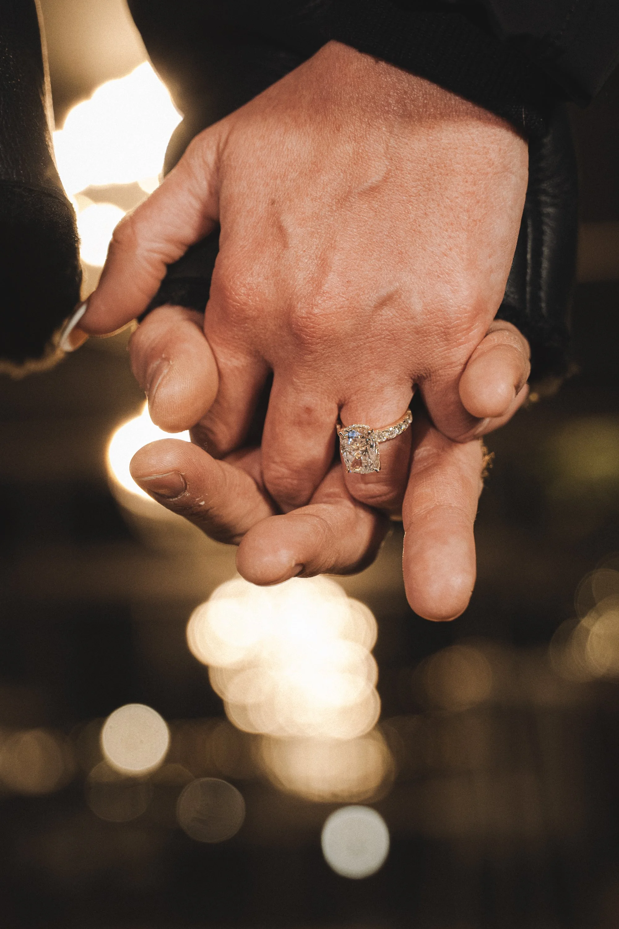 Close-up of two hands holding each other, with a noticeable diamond engagement ring on the ring finger of the hand on top, blurred lights in the background.