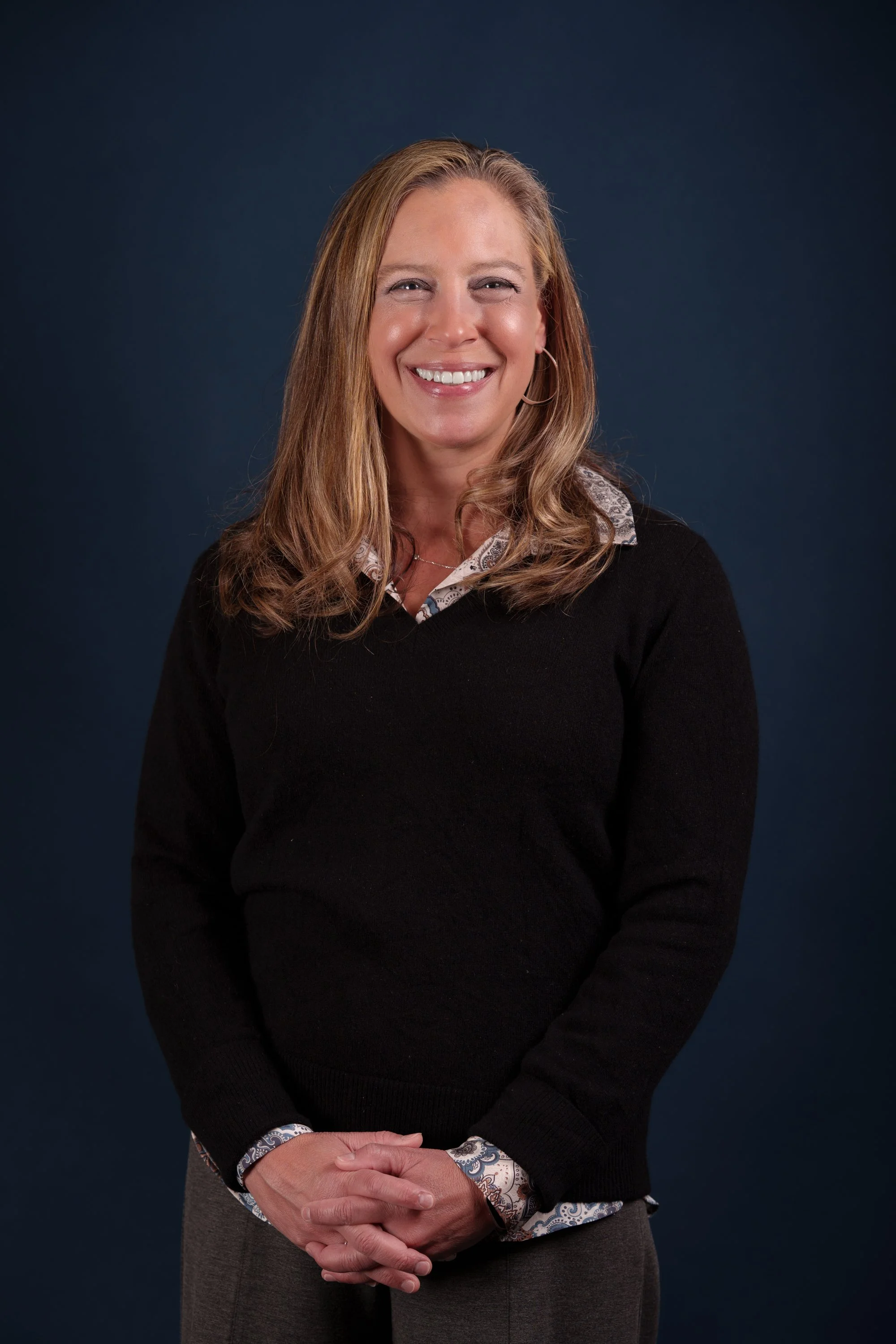 A woman with long blonde hair smiling, wearing a black sweater over a collared shirt with a paisley pattern, standing against a dark background.
