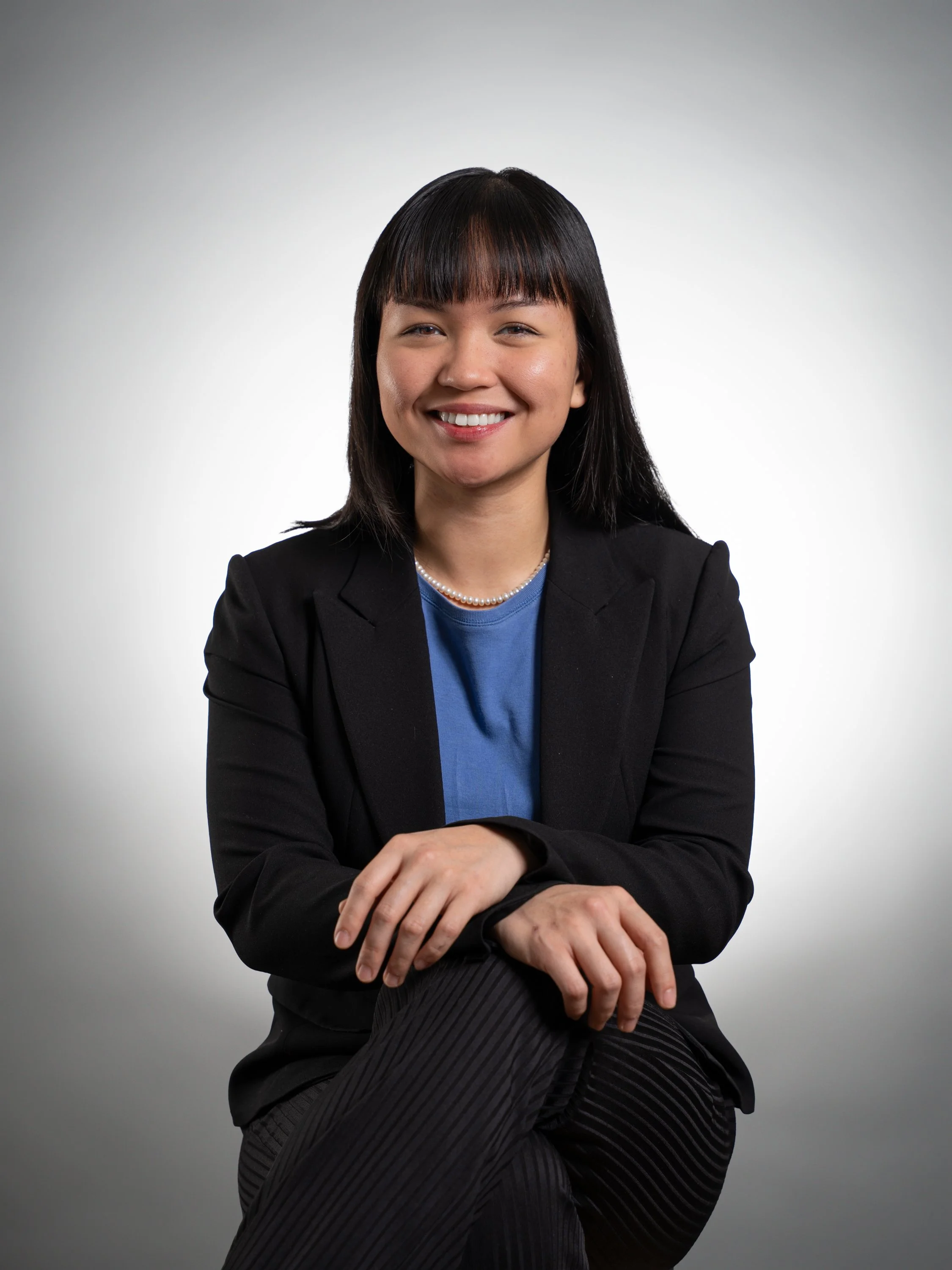 A woman with dark hair and bangs, wearing a black blazer, blue shirt, pearl necklace, and striped pants, smiling with arms crossed in front of a light gradient background.